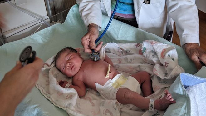 A doctor examines a baby held by a person in a doctor’s office.