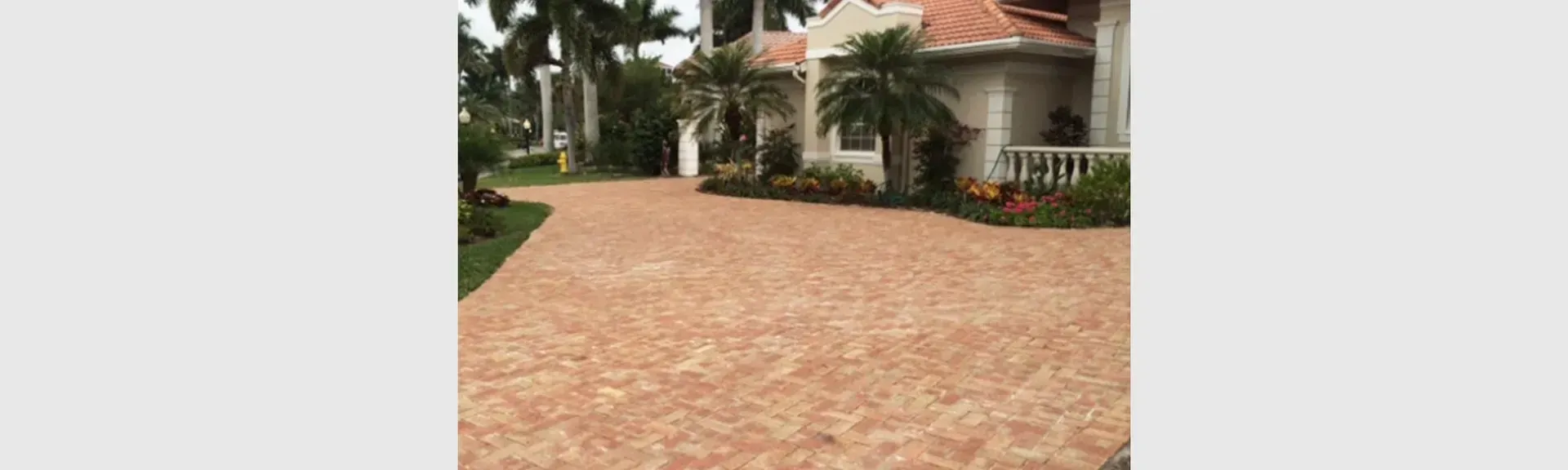 A brick driveway leading to a large house with palm trees.