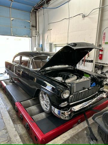 A black car with the hood up is sitting on a dyno in a garage.