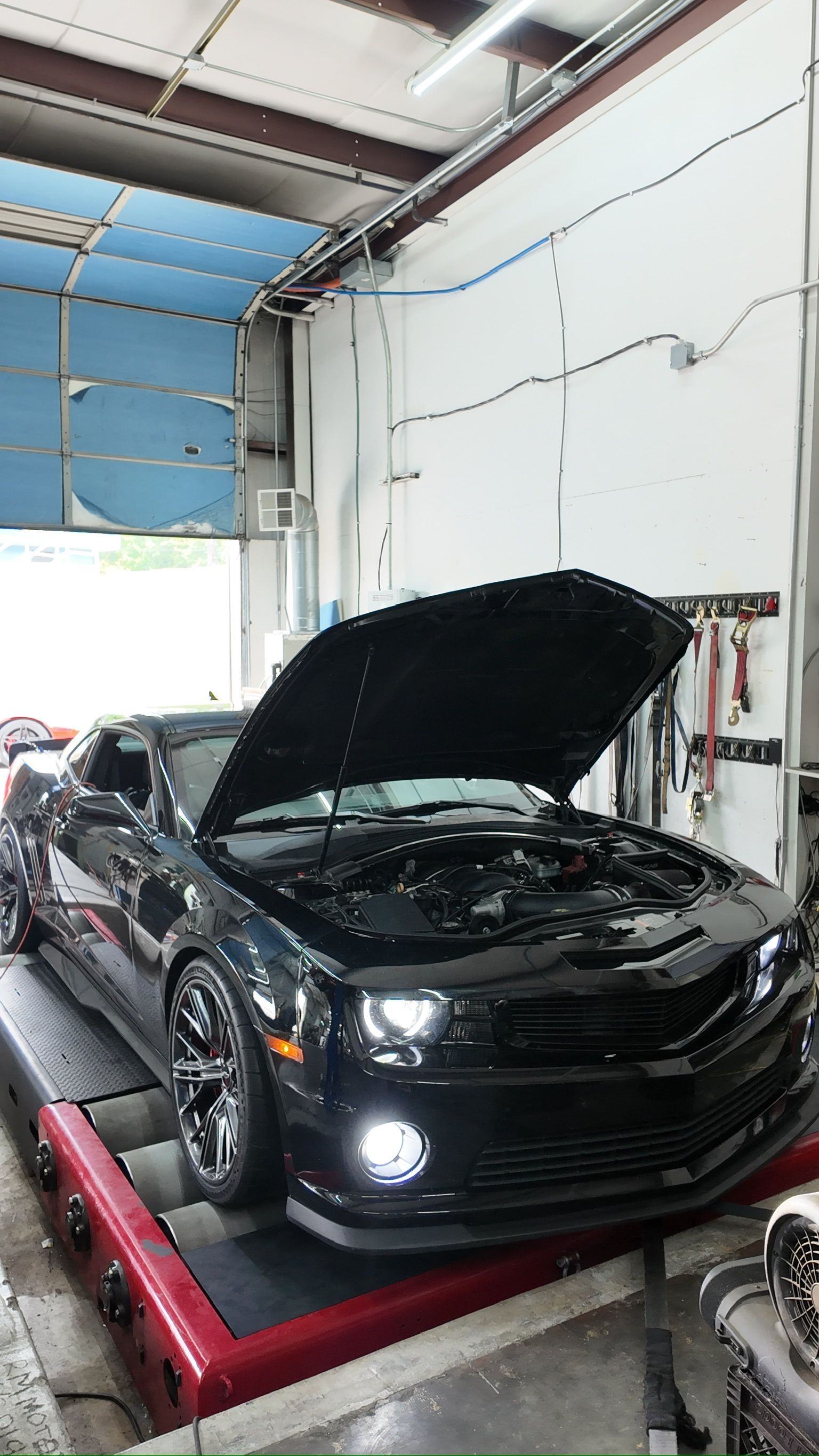 A black car with the hood up is sitting on a dyno in a garage.