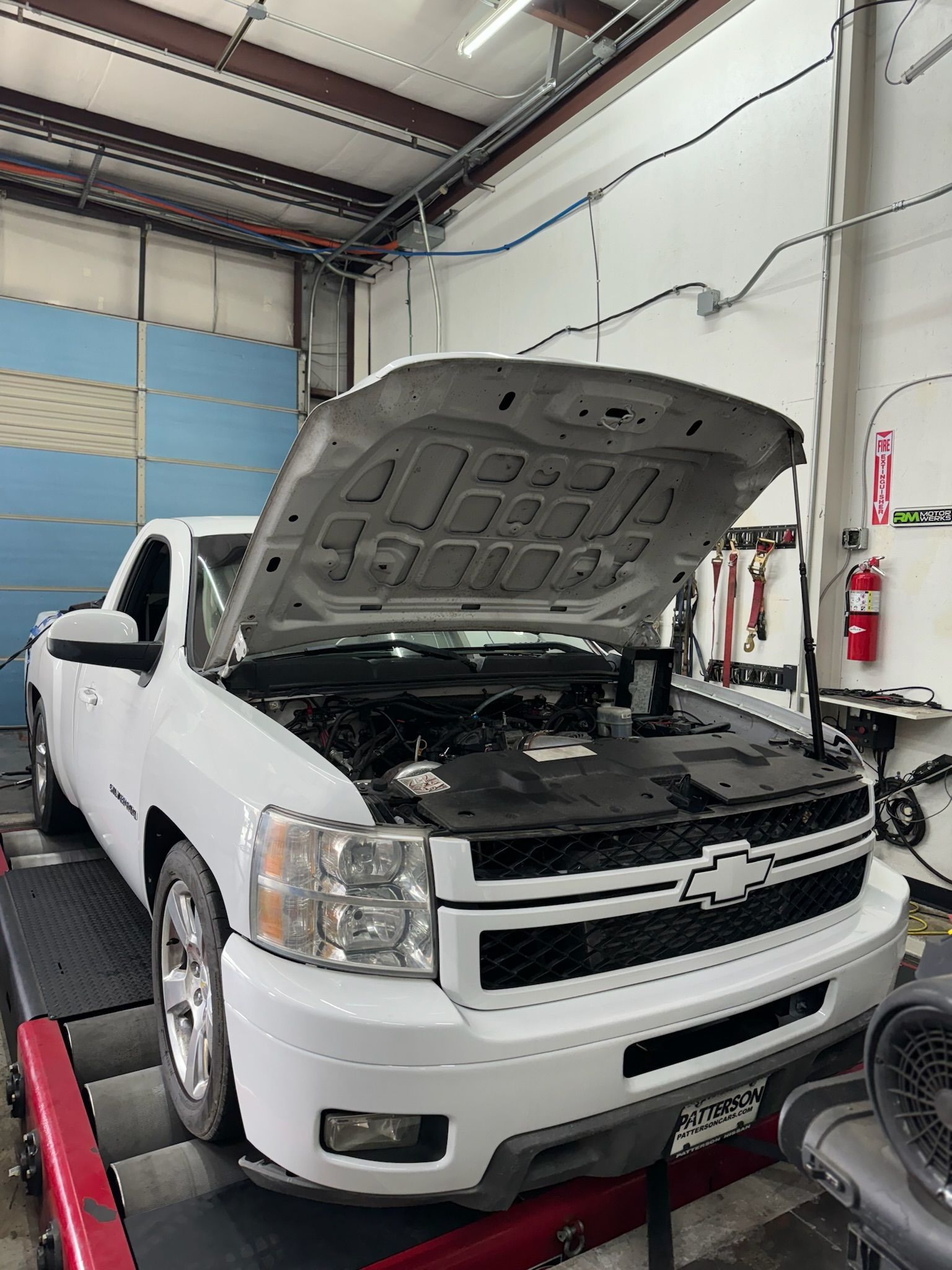 A white truck with the hood up is sitting on a dyno in a garage.