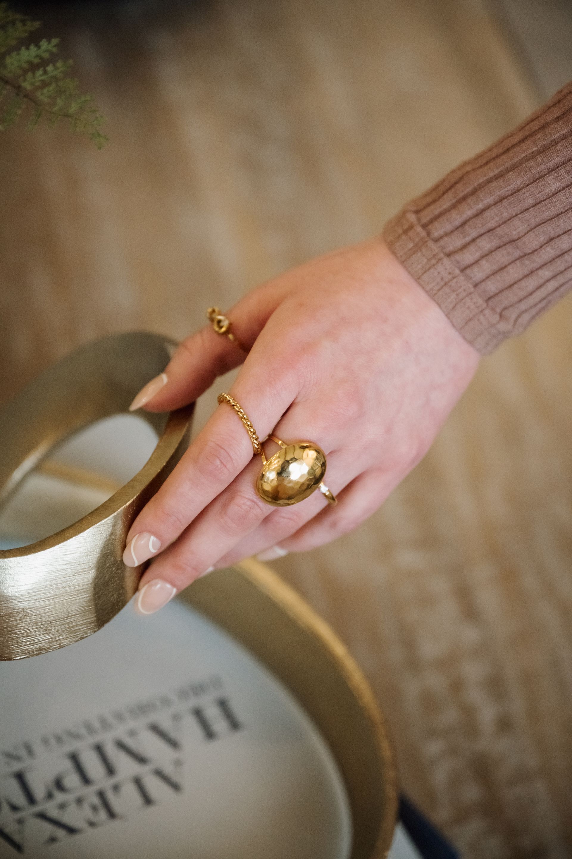 Hand with gold rings reaching toward a gold-toned tray on a carpeted floor
