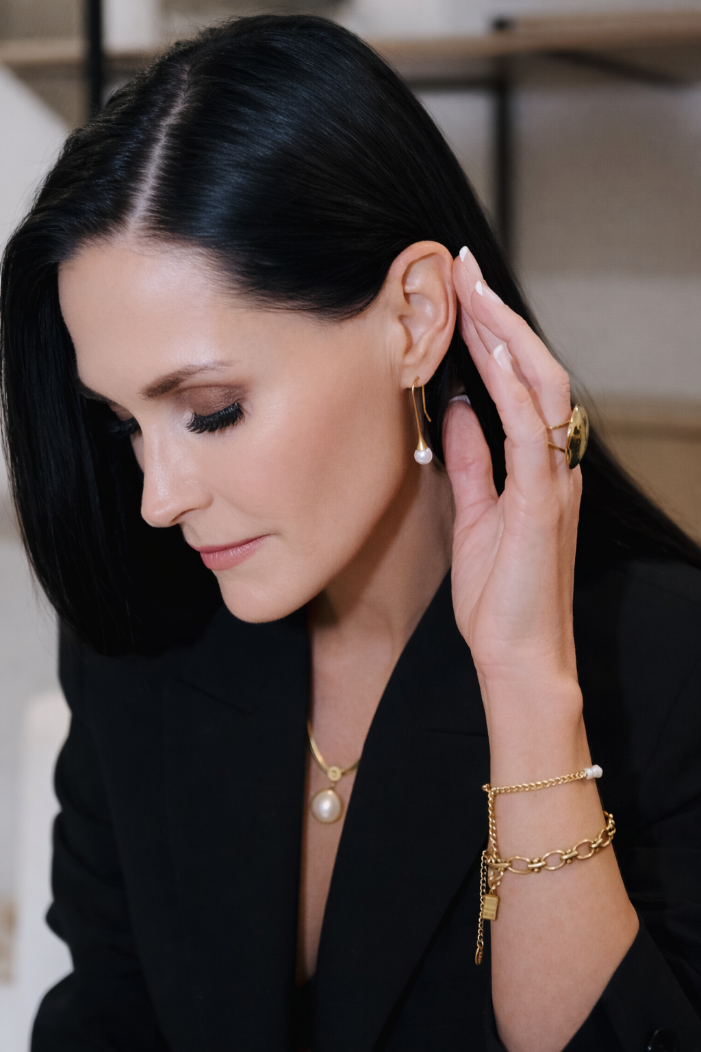 Woman in black outfit adjusting her earring, wearing gold jewelry against a soft indoor background