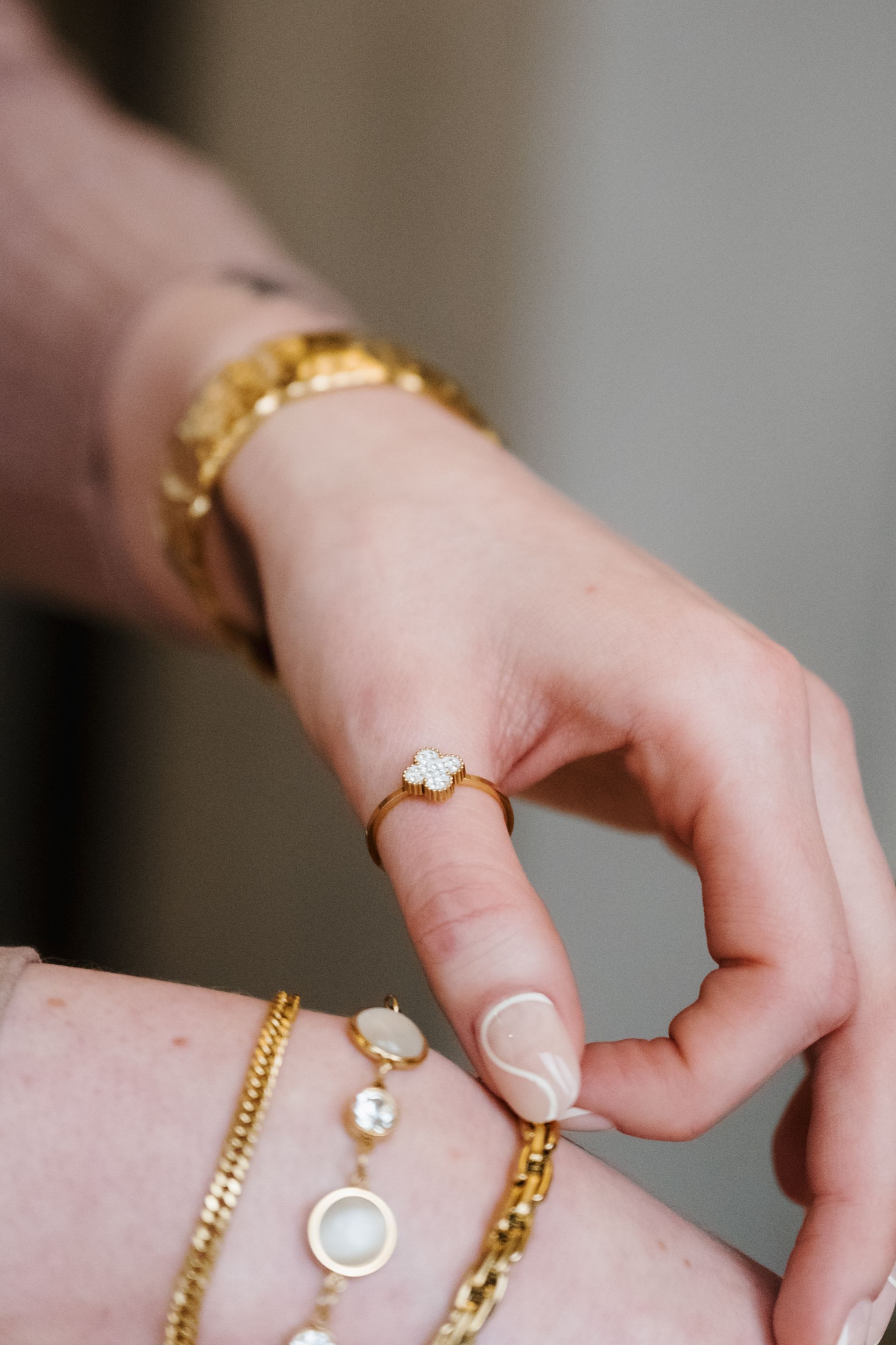 Hands wearing gold jewelry, with a ring and bracelet, held against a soft neutral background