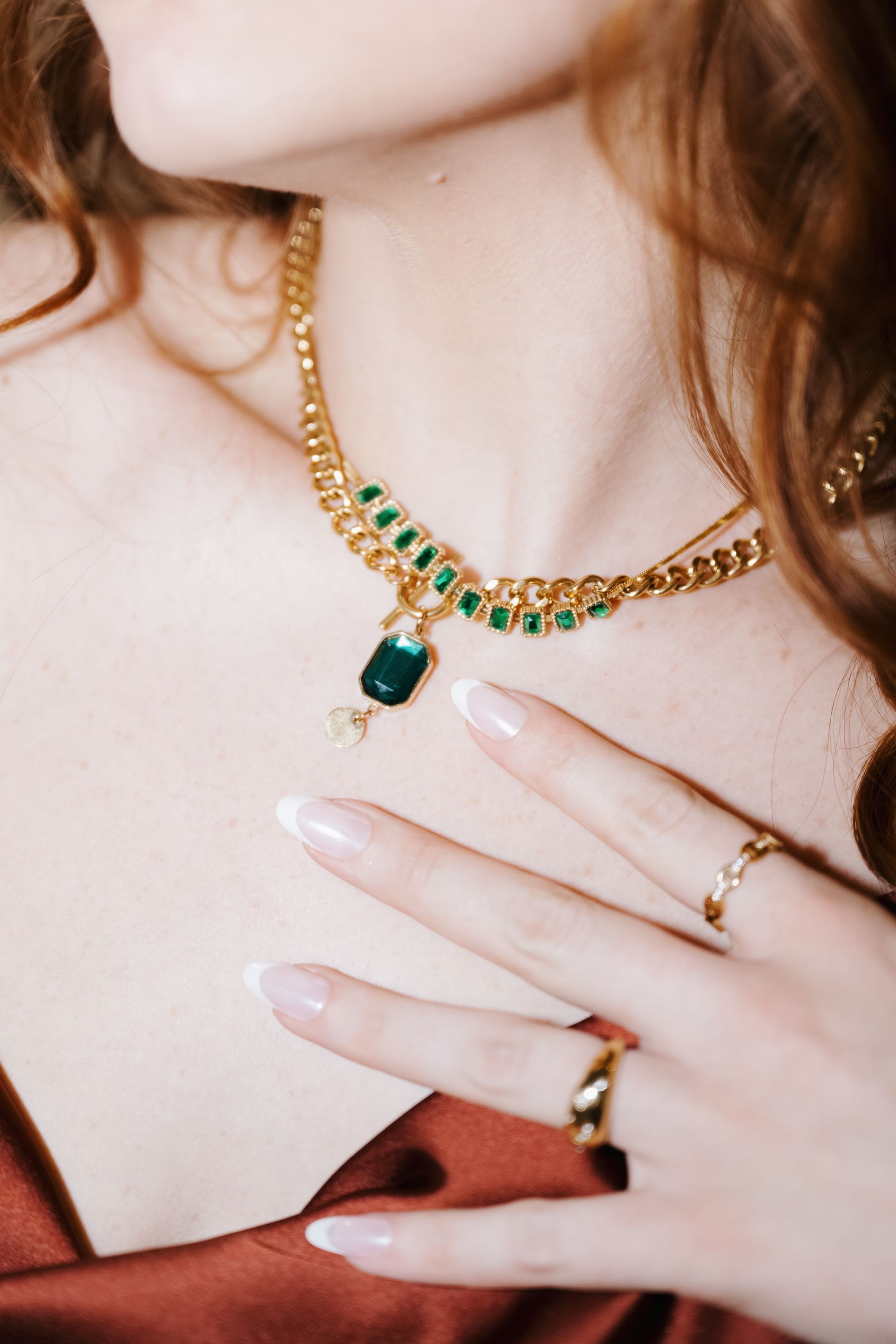 Close-up of a gold necklace with green gemstones resting on a person’s neck and hand with rings.