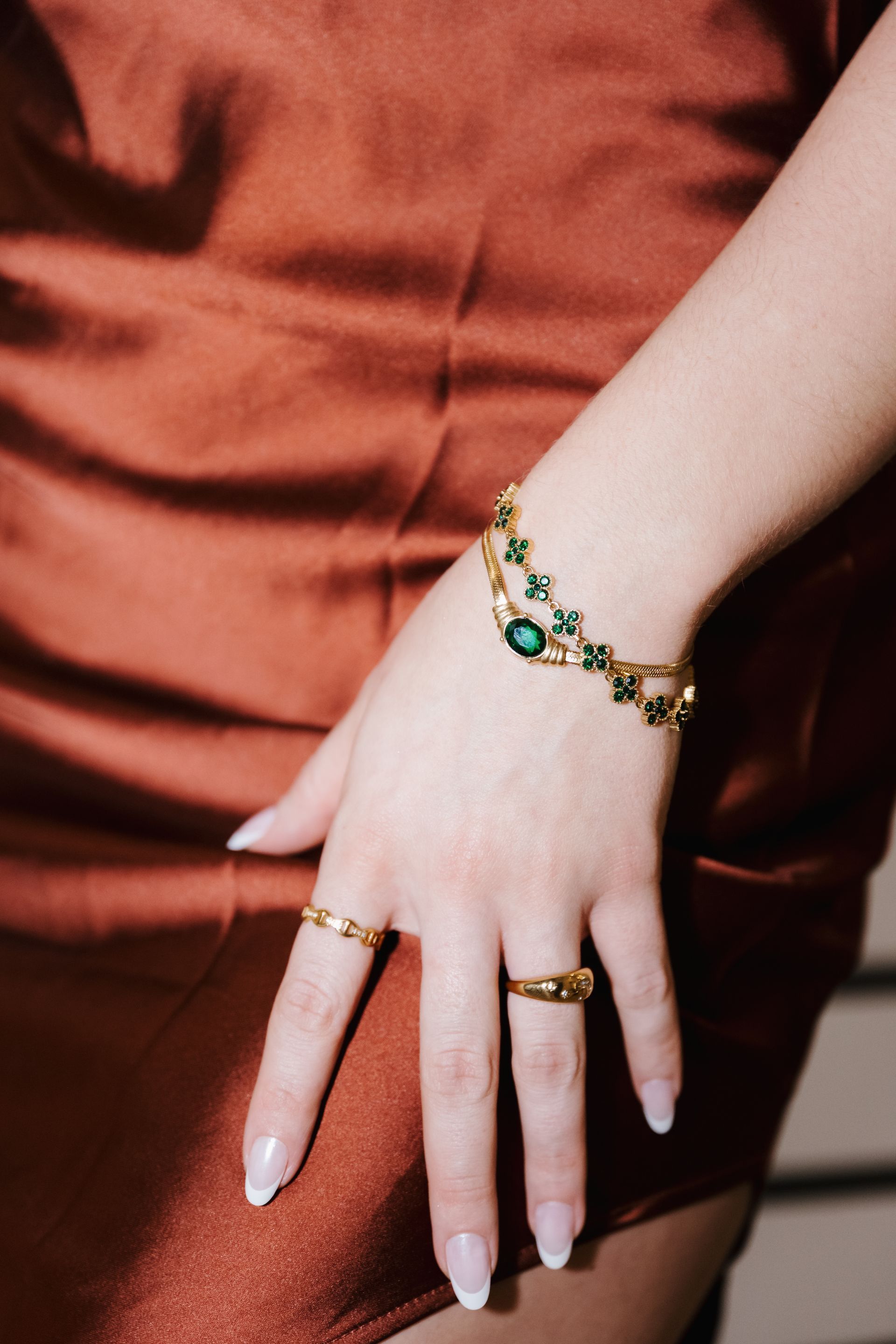 Hand with gold and green jewelry resting on a rust-colored satin dress