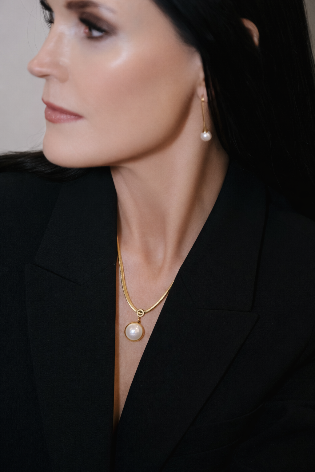Profile portrait of a woman in a black blazer wearing pearl earrings and a gold necklace on a dark background