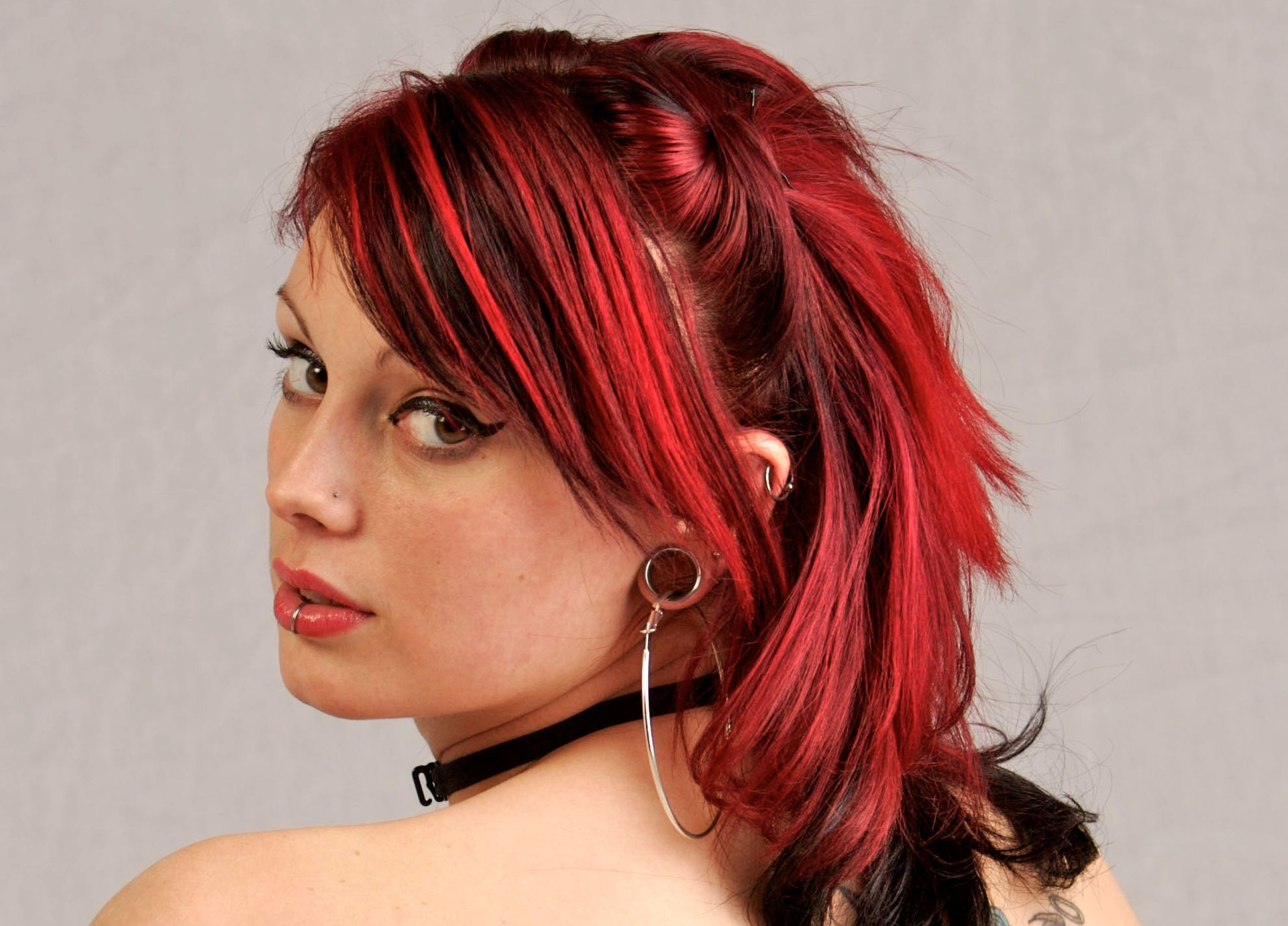 Portrait of a person with red hair and black choker, looking over their shoulder against a plain background