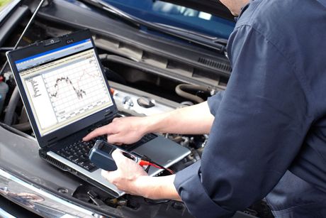 A man is working on a laptop computer under the hood of a car.