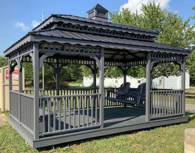Gray gazebo with a dark roof and two wooden swing seats, located in a grassy area.