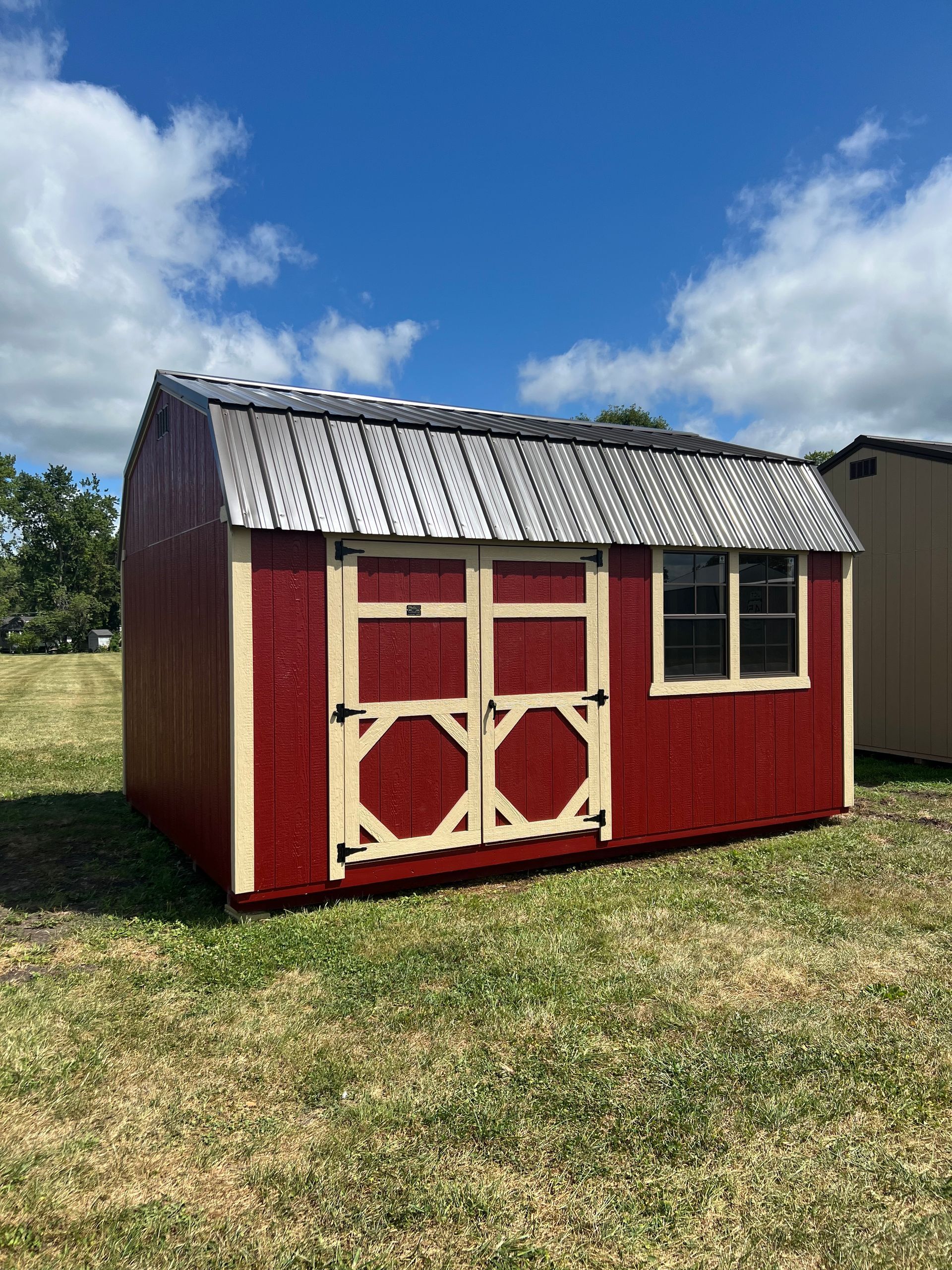 Red shed with tan trim, small window, and wood shingle roof on grassy field under a blue sky.