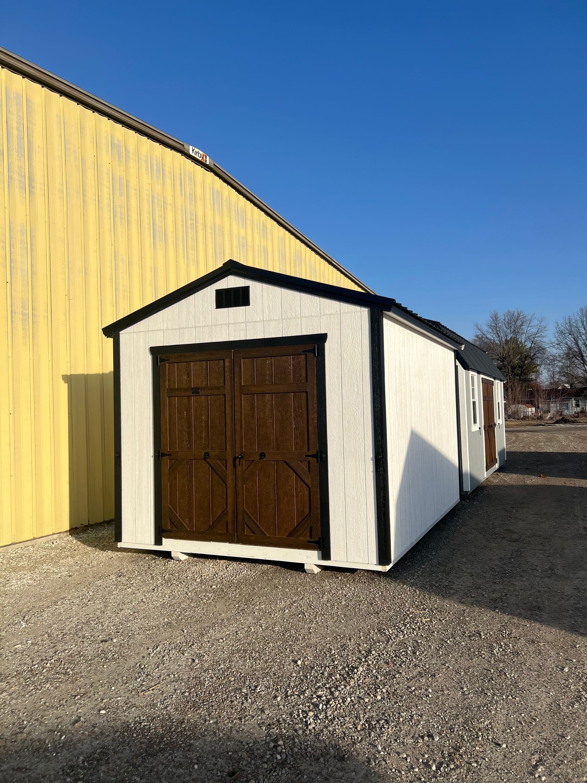 White shed with brown double doors, black trim, adjacent to a yellow building, gravel ground.