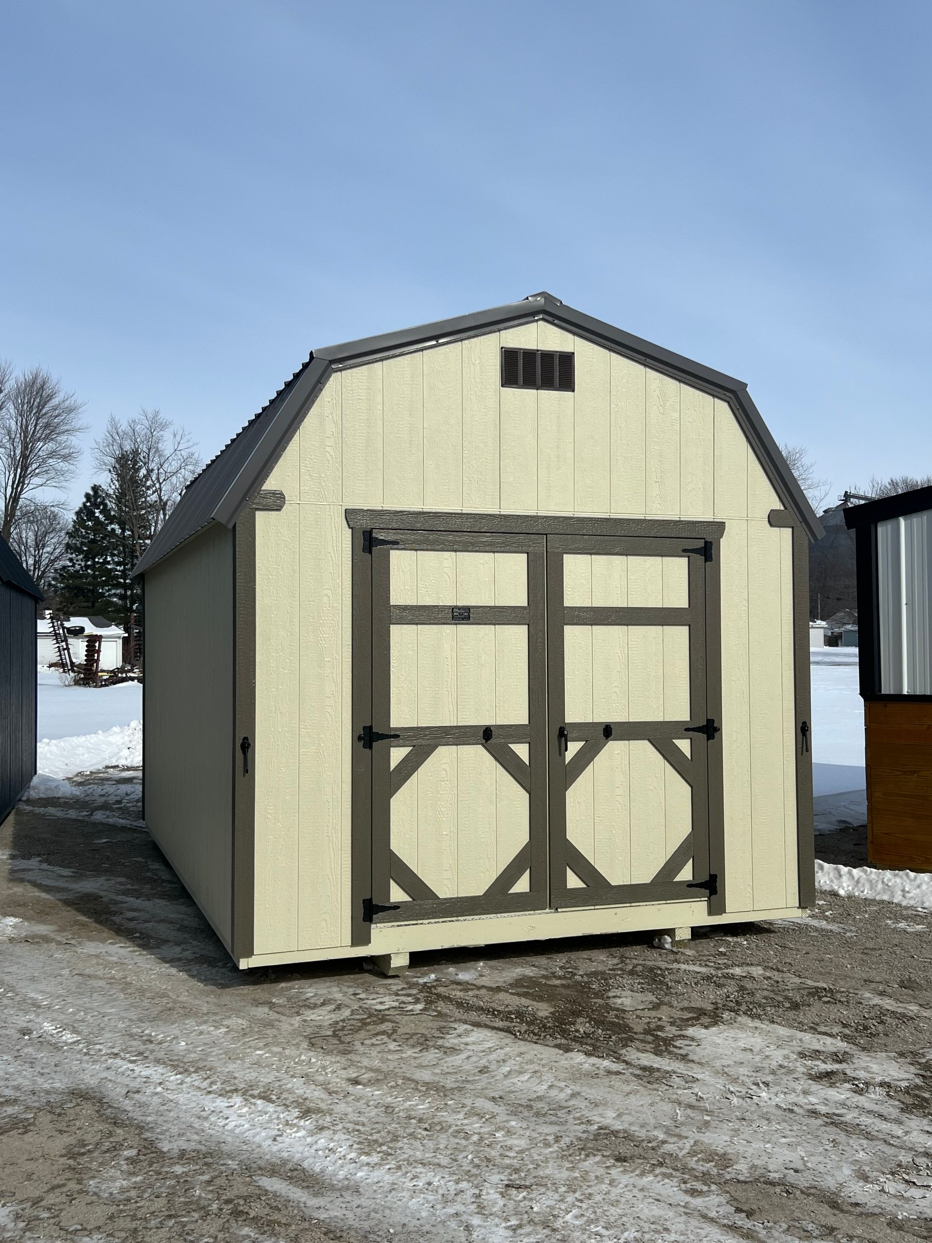 Tan barn-shaped shed with brown trim, double doors, and a snow-covered ground.