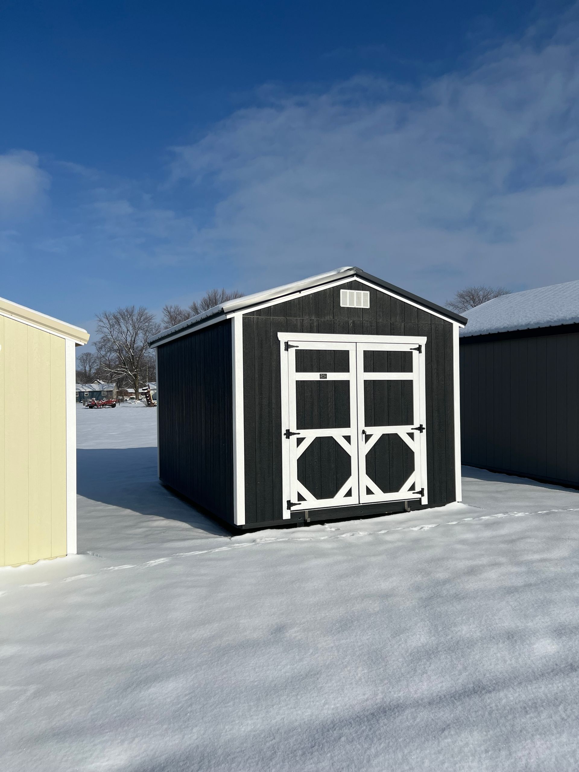 Dark grey shed with white trim, double doors, in a snowy field on a sunny day.