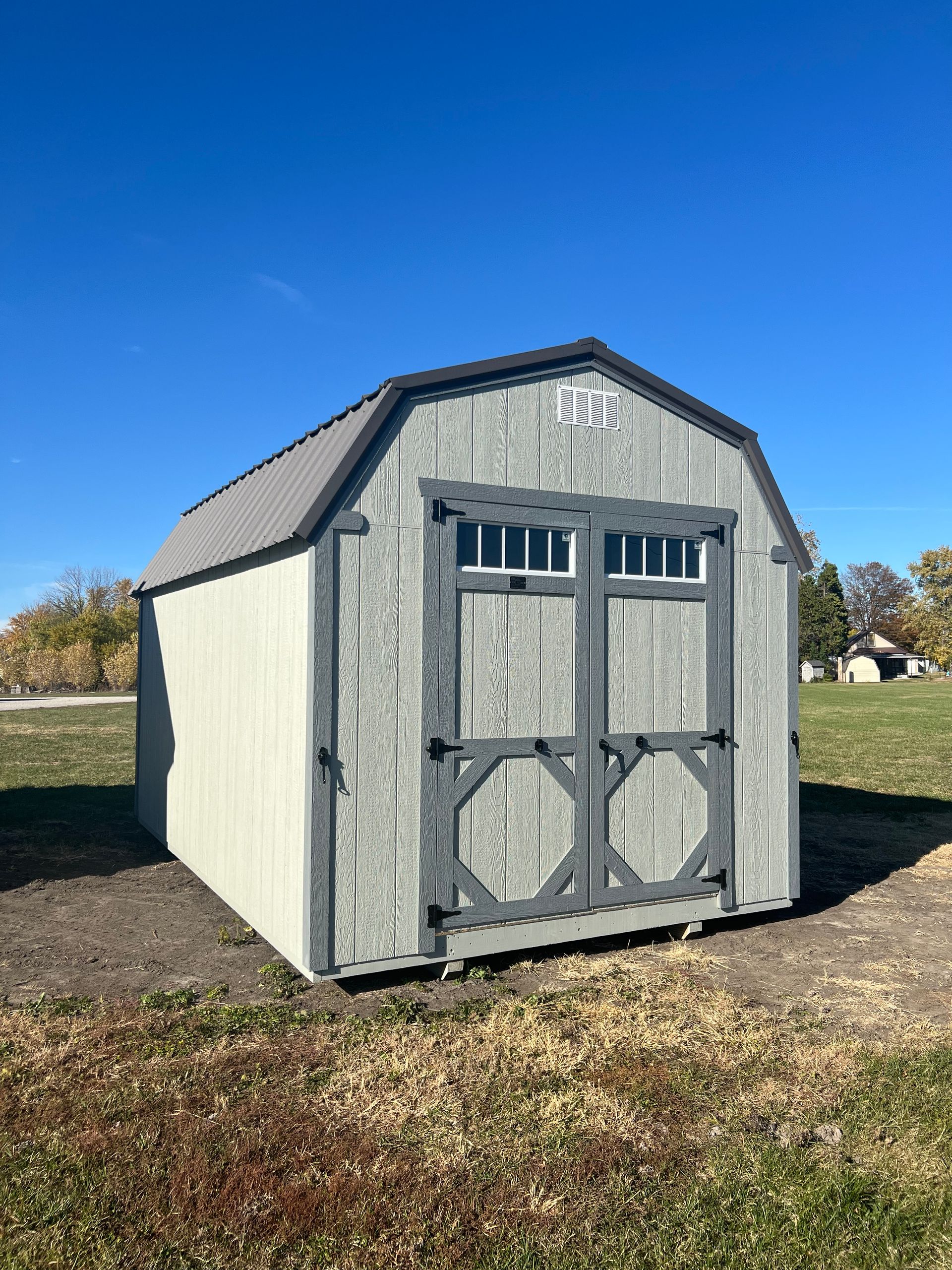 Gray shed with barn-style doors and a dark roof, on a grassy field under a blue sky.