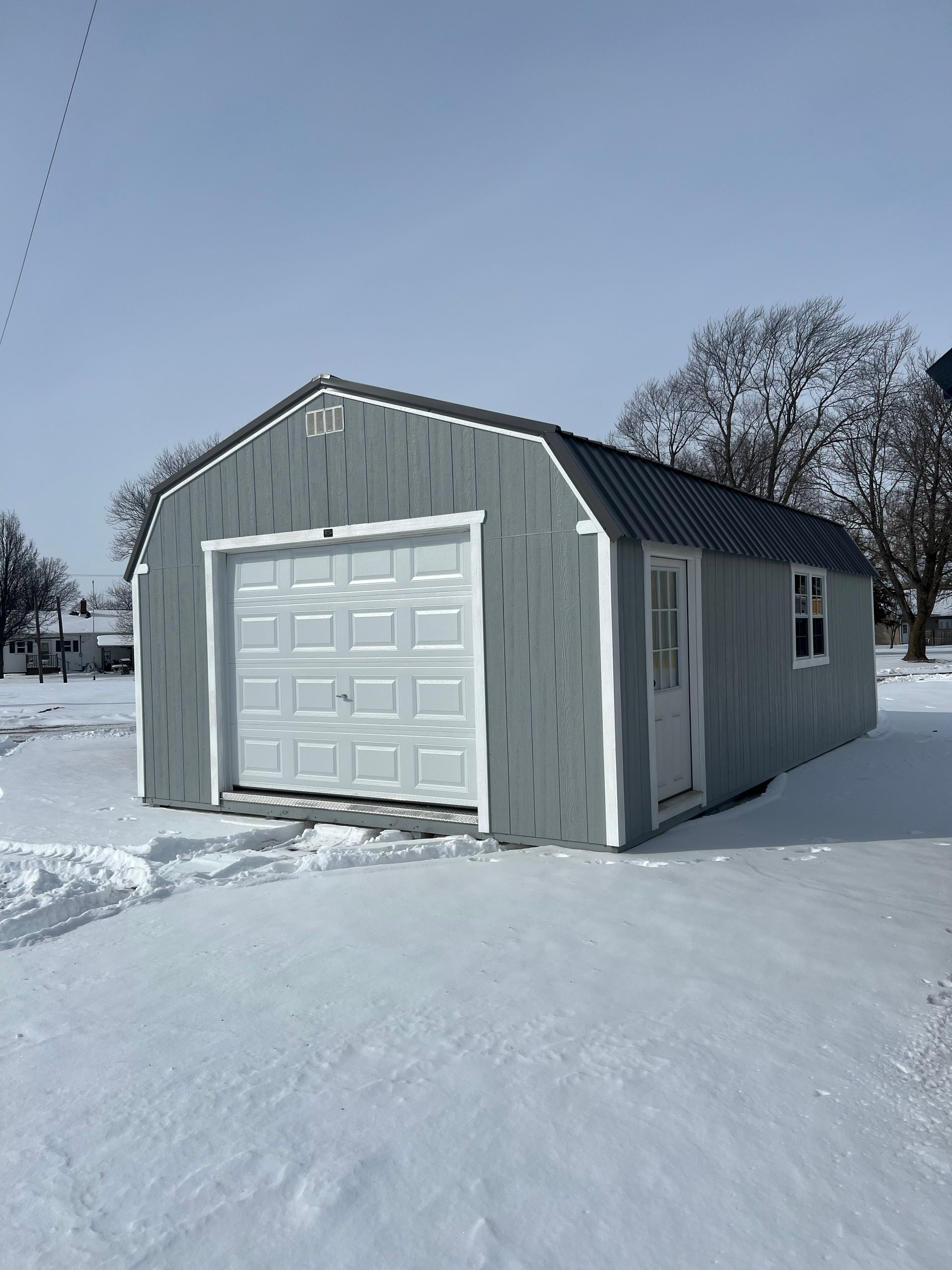 Gray shed with white garage door and snow-covered ground under a clear sky.