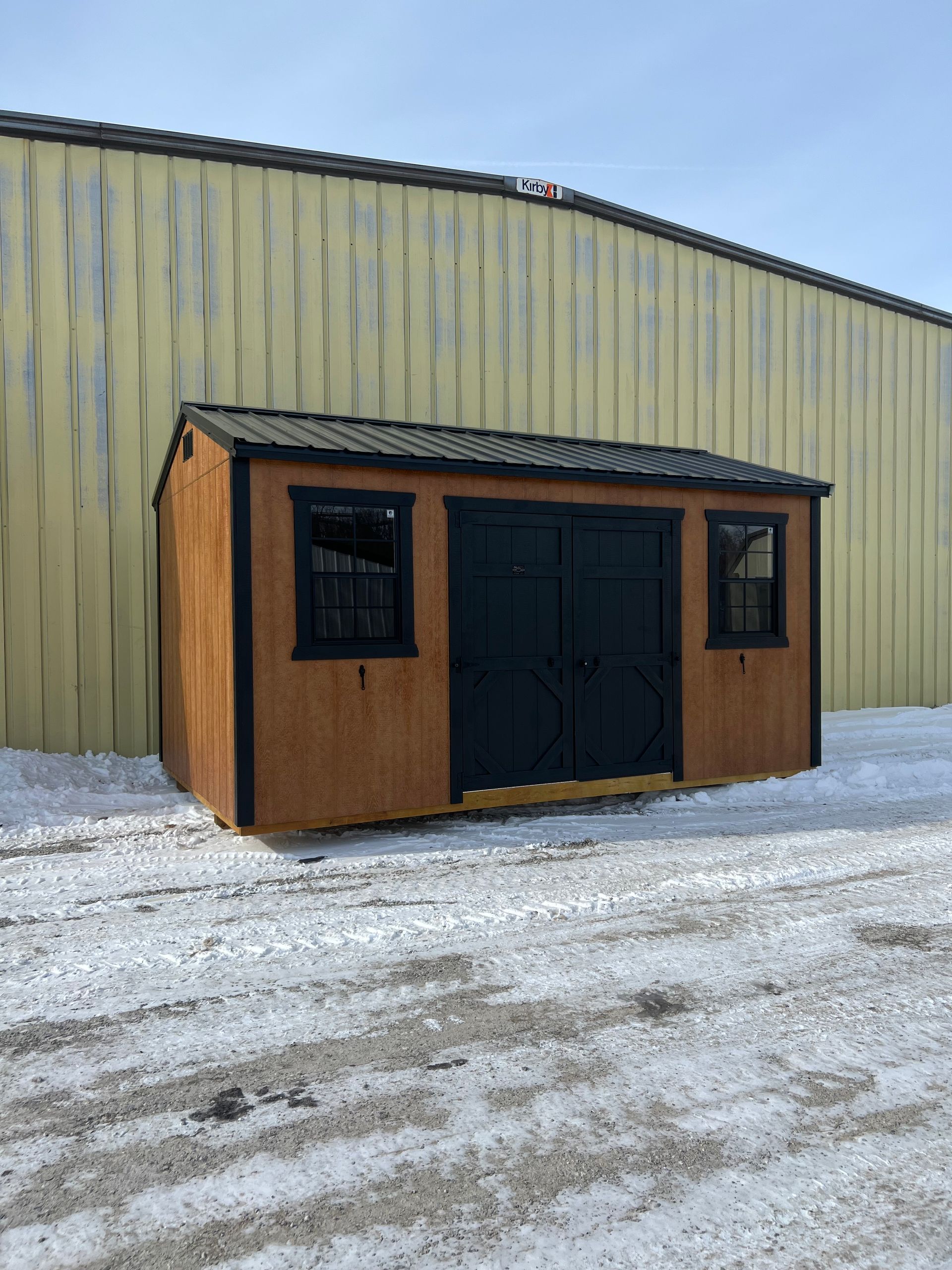 Tan shed with black doors and trim, two windows, set in snow, against a yellow building.