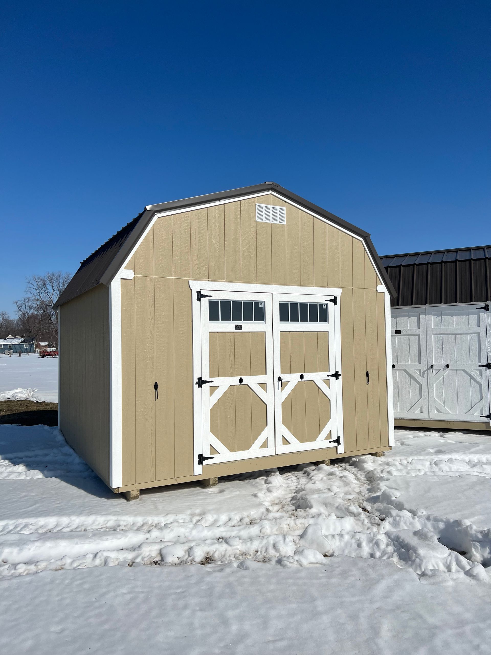 Tan barn-style shed with white trim and double doors on snow, under a clear blue sky.