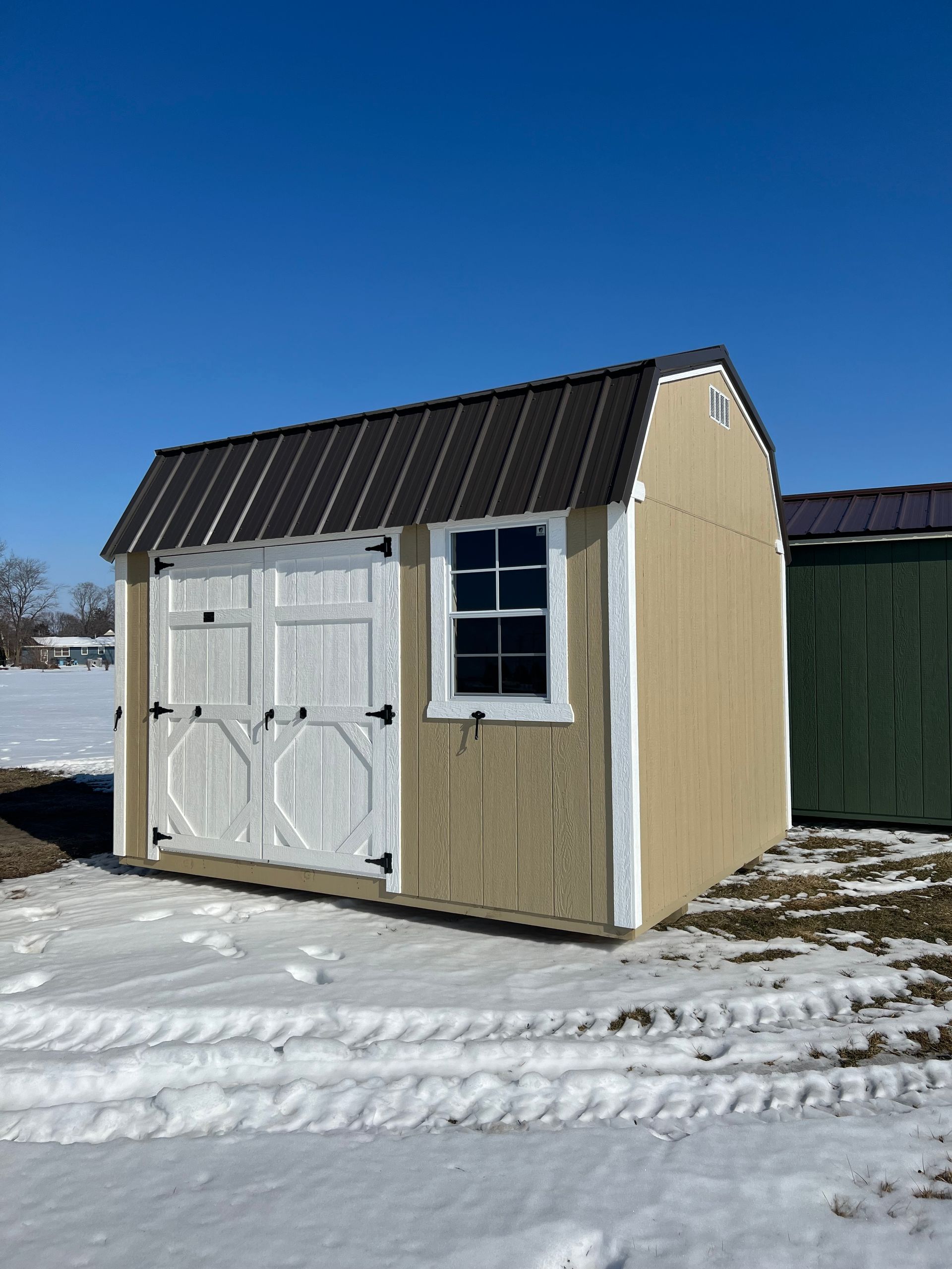 Tan shed with white doors, window, and trim, brown roof, snowy ground, and blue sky.