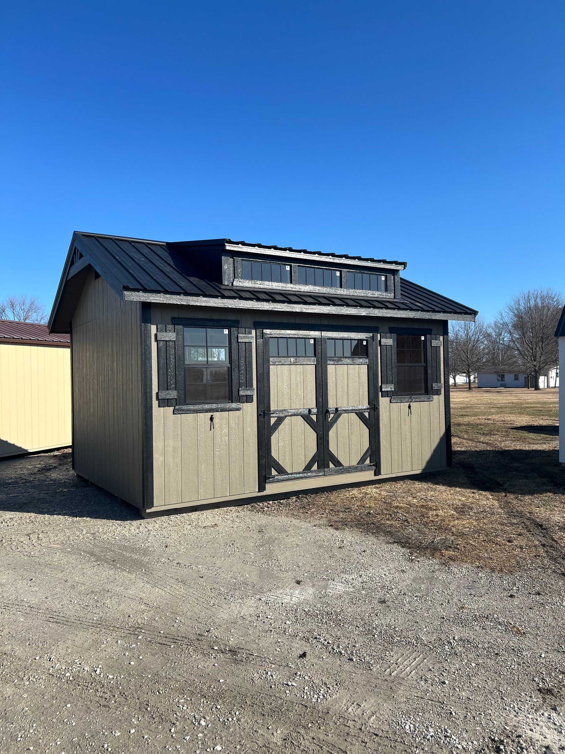 Tan shed with black roof and trim on a gravel lot under a blue sky.