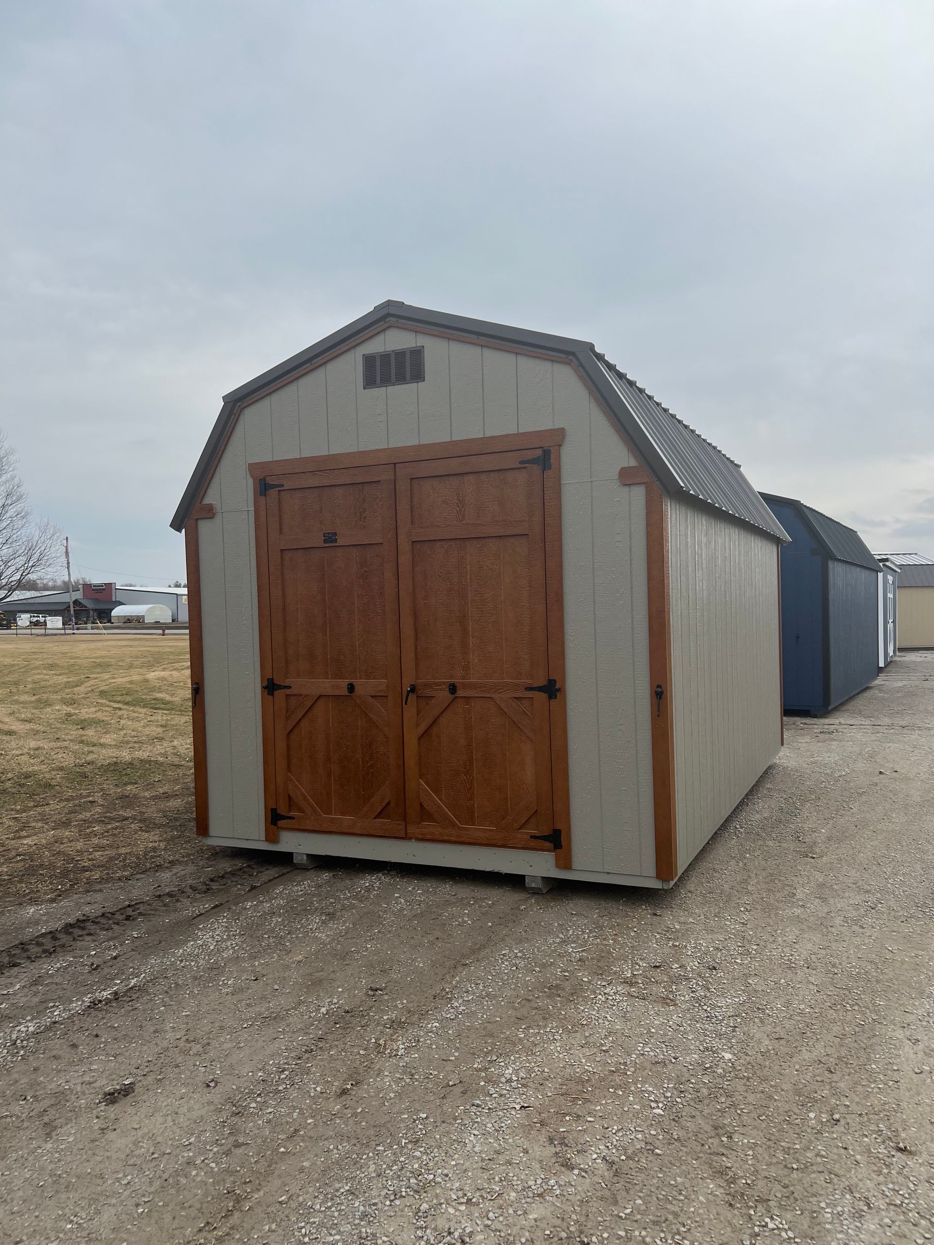 Tan shed with brown doors, gravel ground, and blue shed in the background.