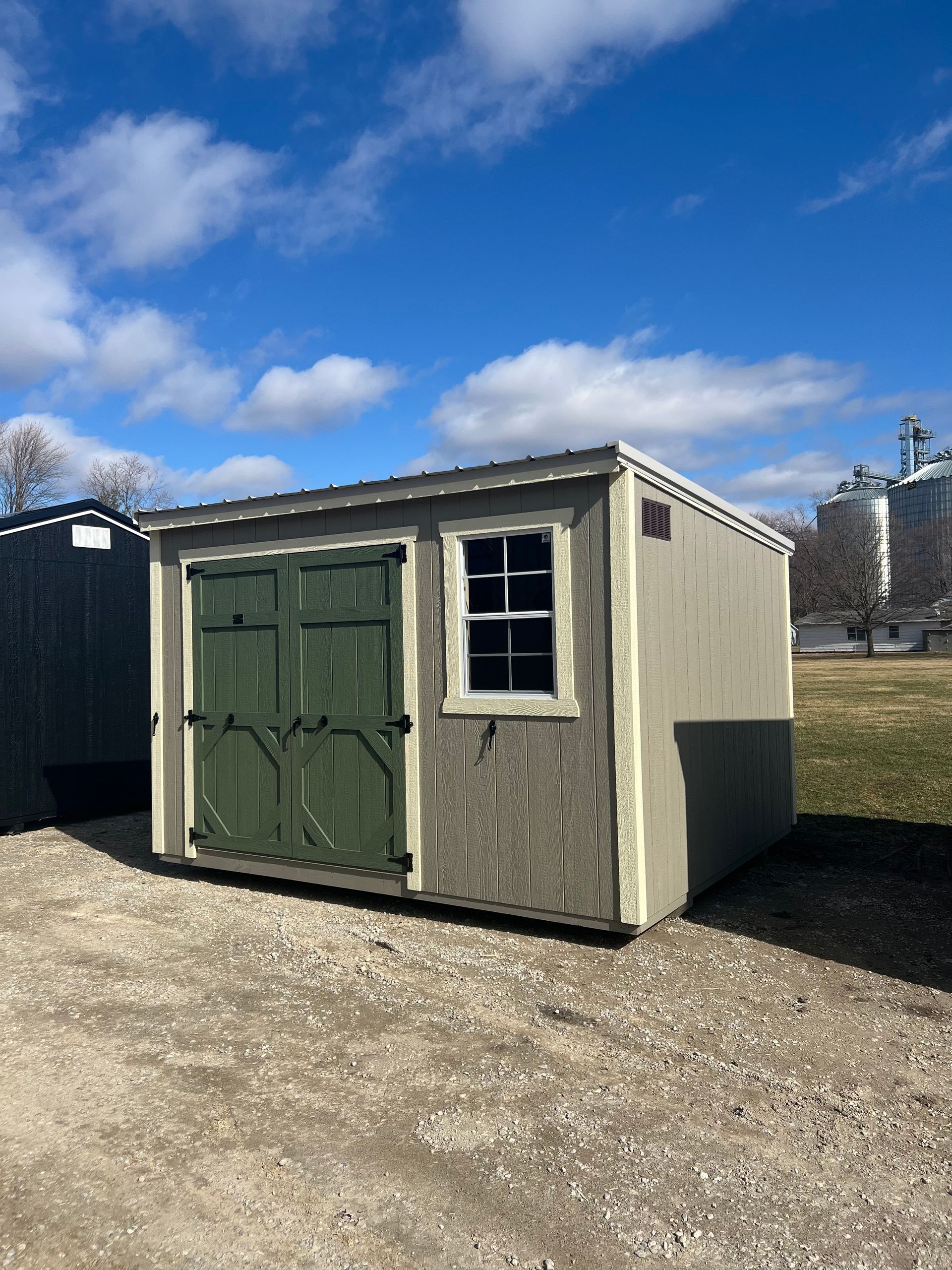 Tan shed with green doors and window against a blue sky with clouds.