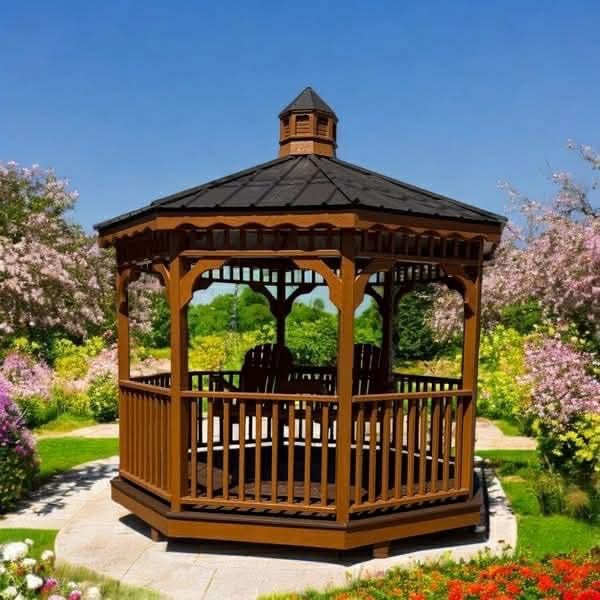 Wooden gazebo in a garden with chairs, surrounded by flowers and trees under a blue sky.