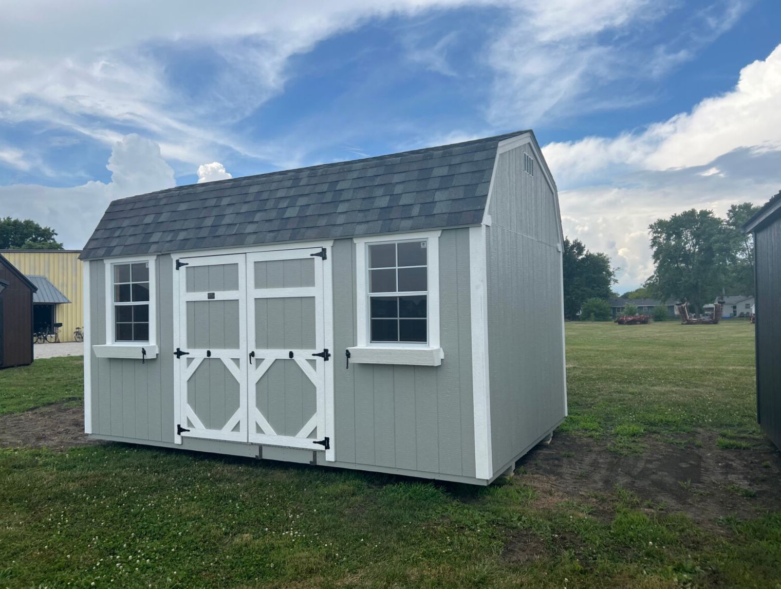 A gray barn-style storage shed with white trim, double doors, and two windows with flower boxes, standing in a field.