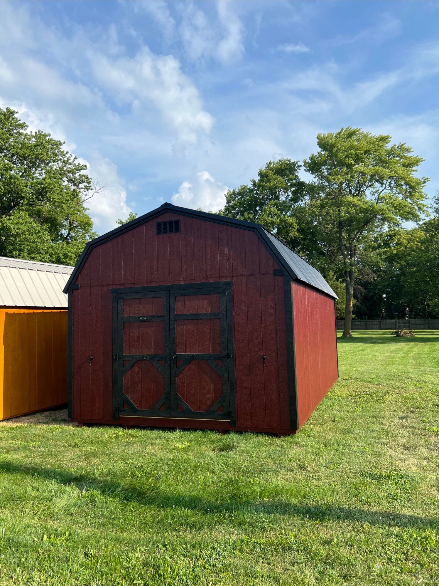 A red wooden barn-style shed with black trim and double doors stands in a grassy field under a sunny blue sky.