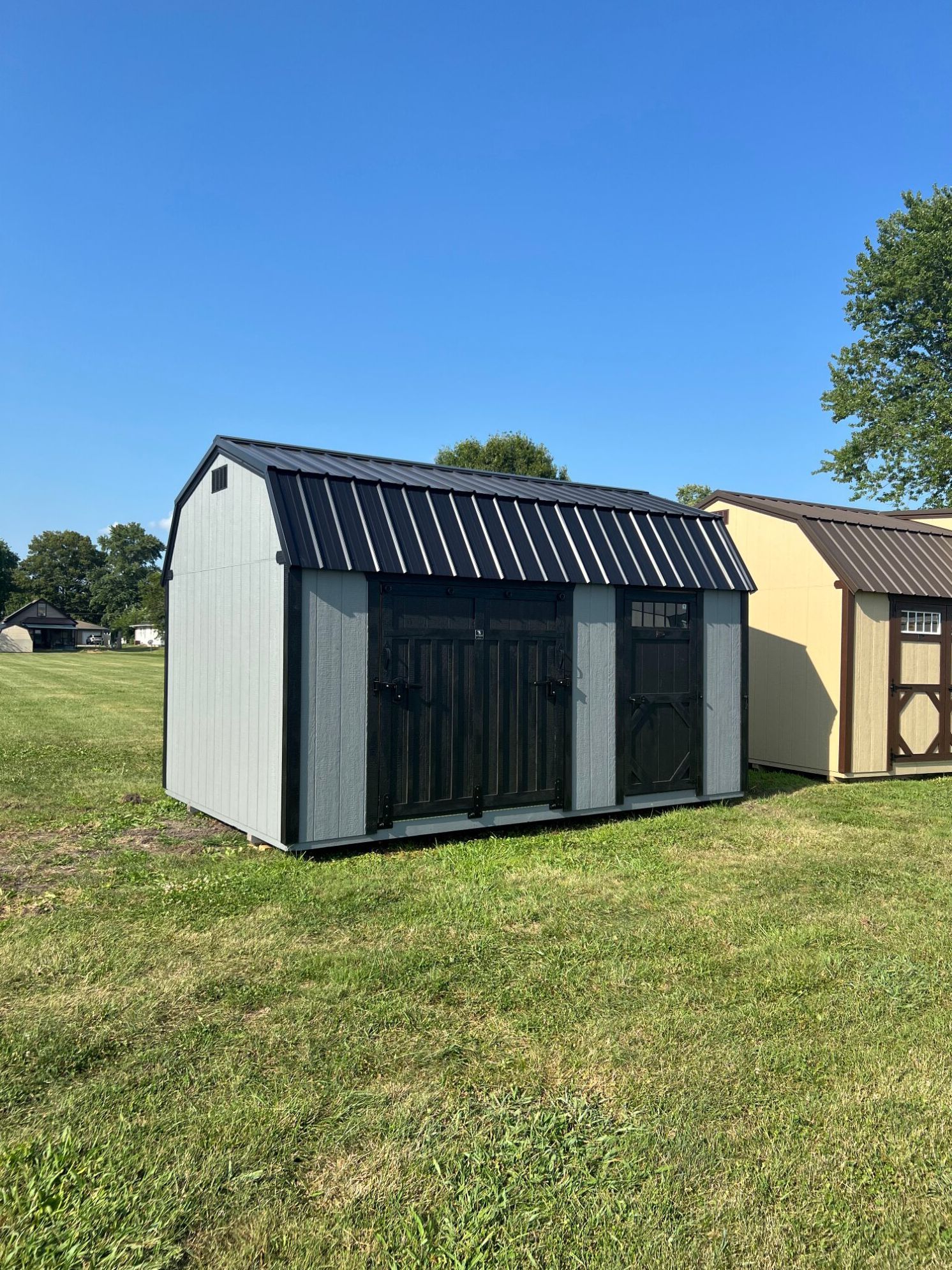 A light gray shed with a dark metal gambrel roof and black doors sits on a grassy field under a clear blue sky.