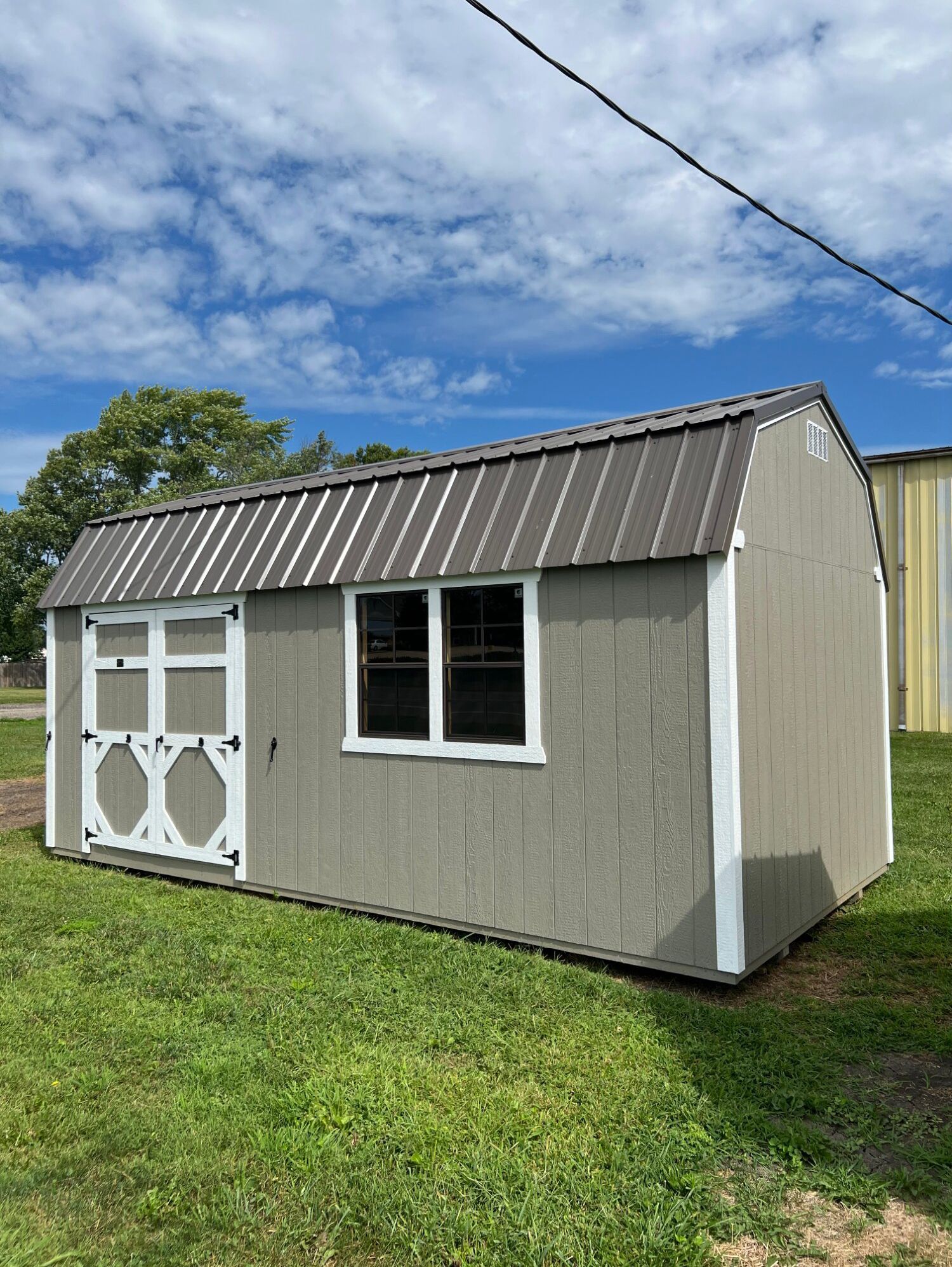 A gray, wooden storage shed with a dark metal roof, white trim, double doors, and two windows in a grassy field.
