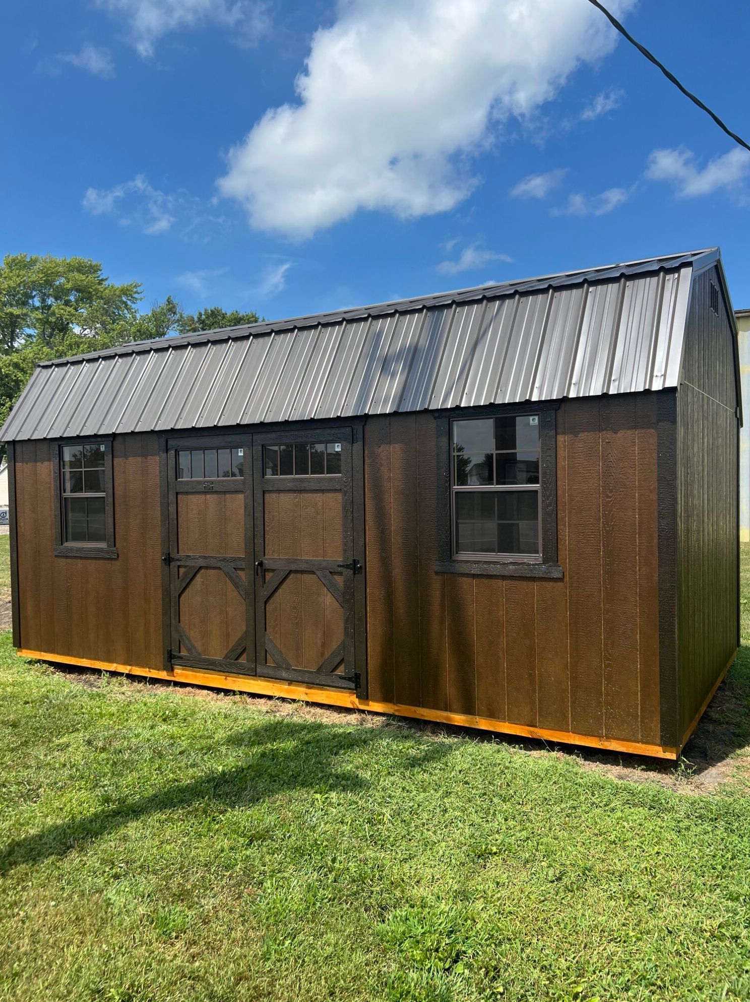 A brown shed with a dark metal roof, double doors, and two windows, positioned on a grassy lawn under a blue sky.