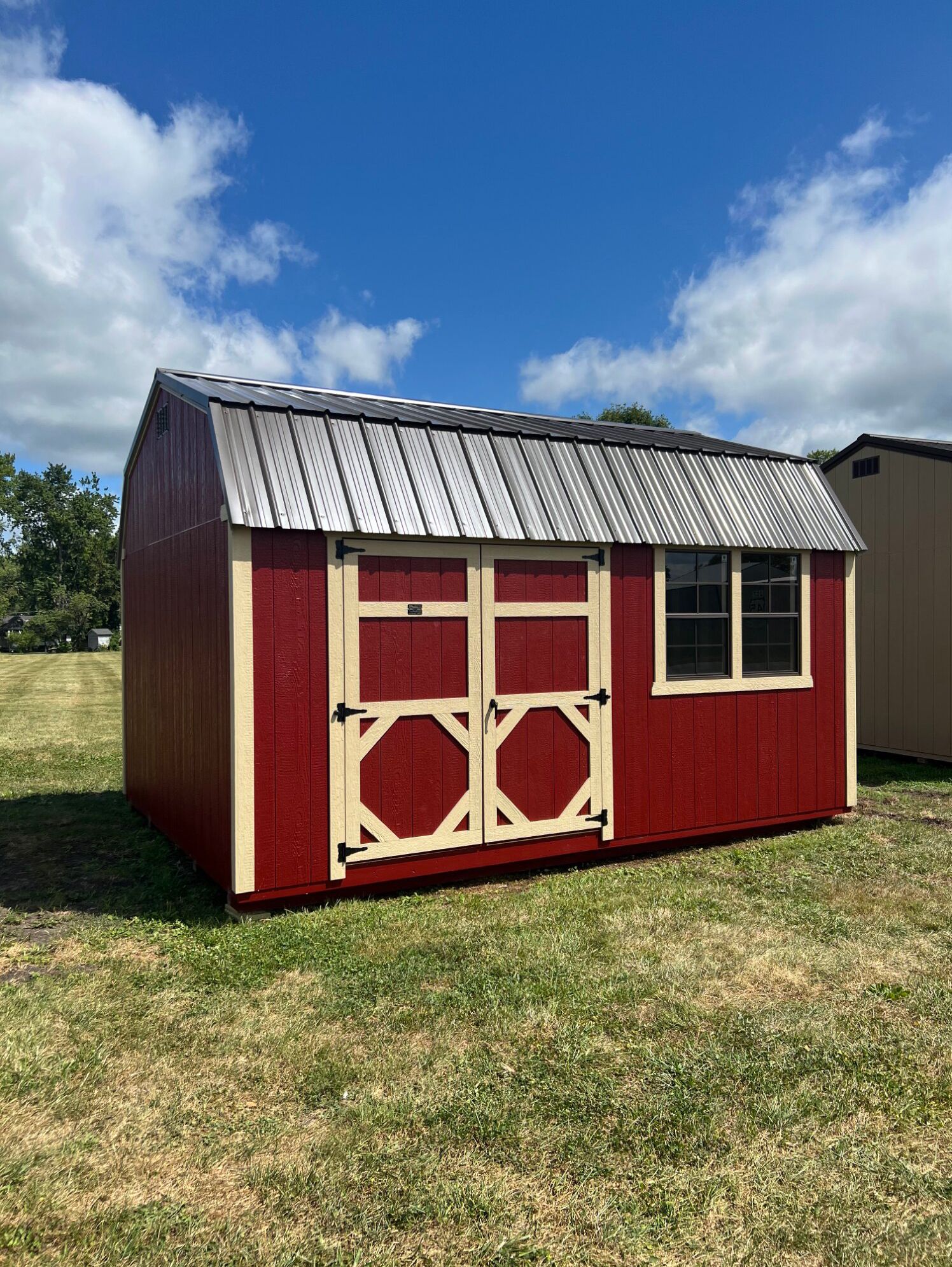 A red and beige backyard shed with a silver metal roof, standing in a grassy field under a blue sky with clouds.