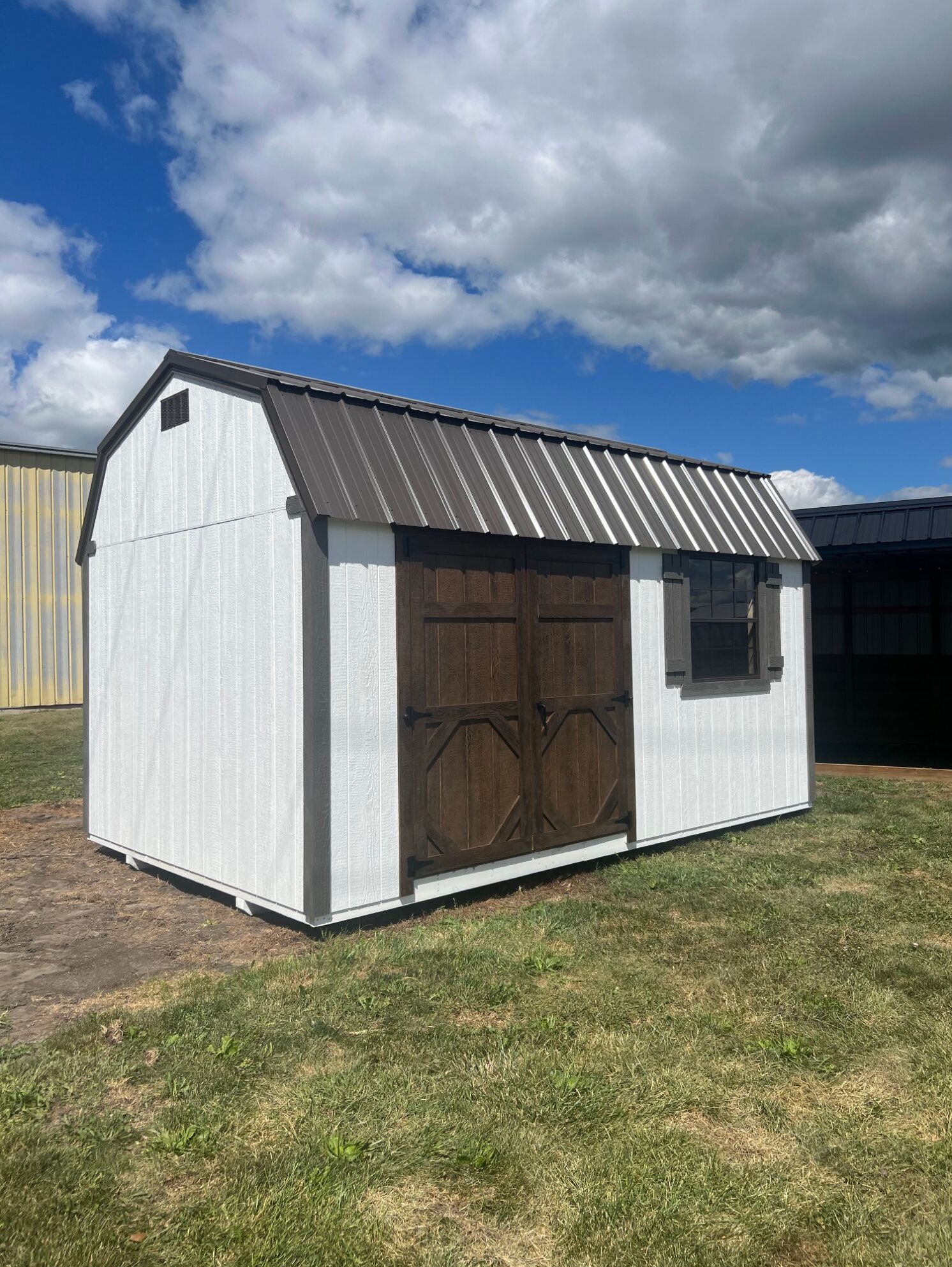 A white barn-style shed with a brown metal roof, dark brown double doors, and a window, sitting on grass under a blue sky.
