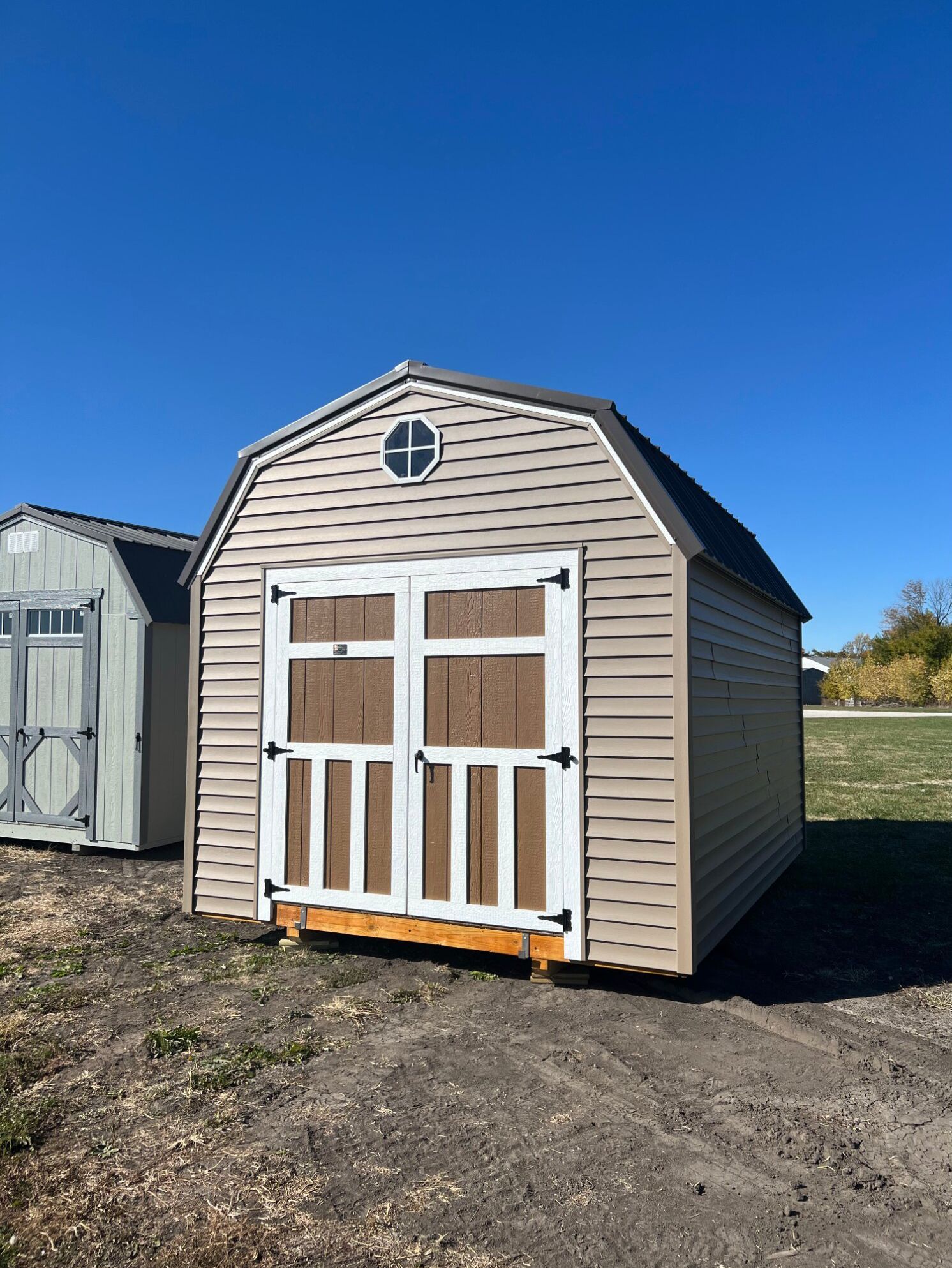 A beige, gambrel-roof storage shed with white-trimmed double doors and a small circular window under a clear blue sky.
