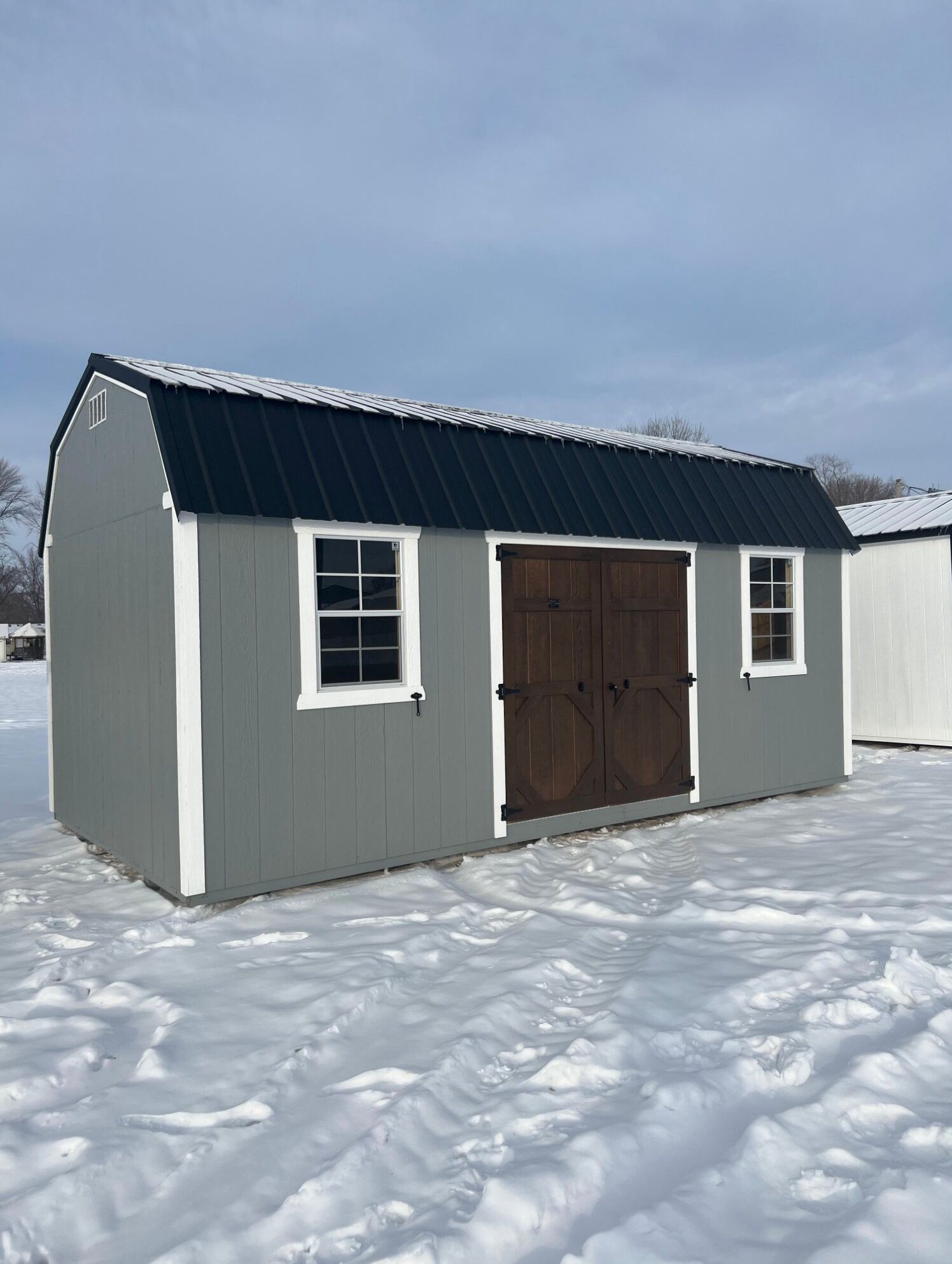 A gray storage shed with a black metal gambrel roof, white trim, two windows, and double wooden doors, set in the snow.