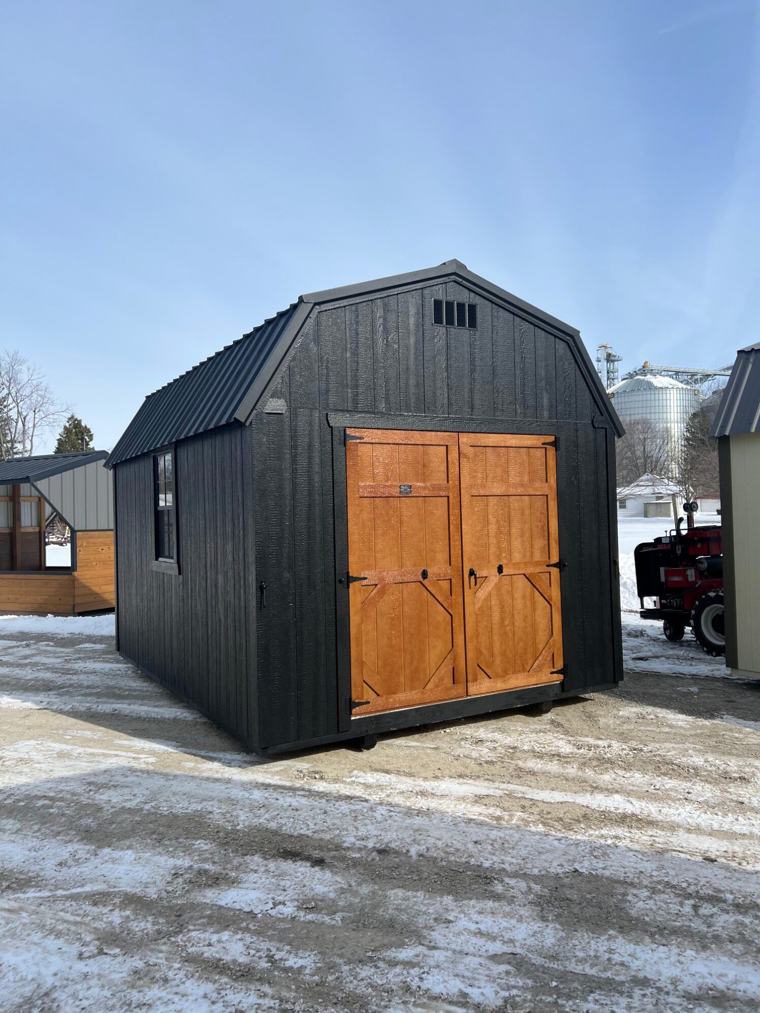 A black, gambrel-roof shed with natural wood double doors, standing on a snow-covered gravel lot under a bright blue sky.