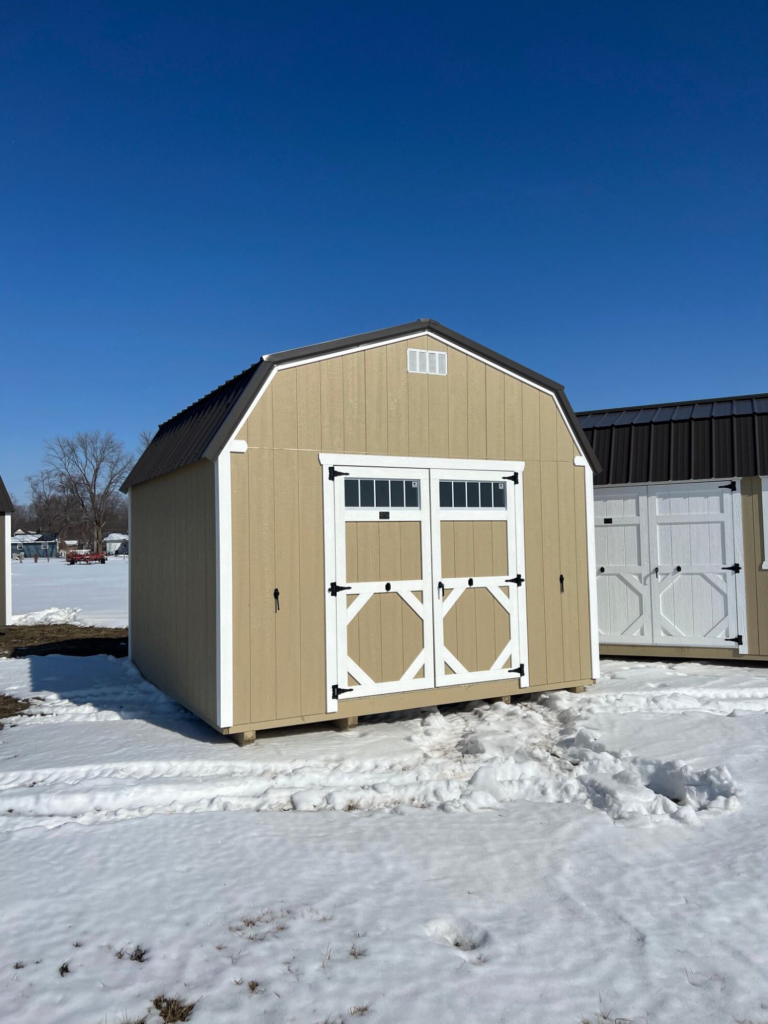 A beige gambrel-roof shed with white trim and double doors sitting on snow-covered ground under a clear blue sky.