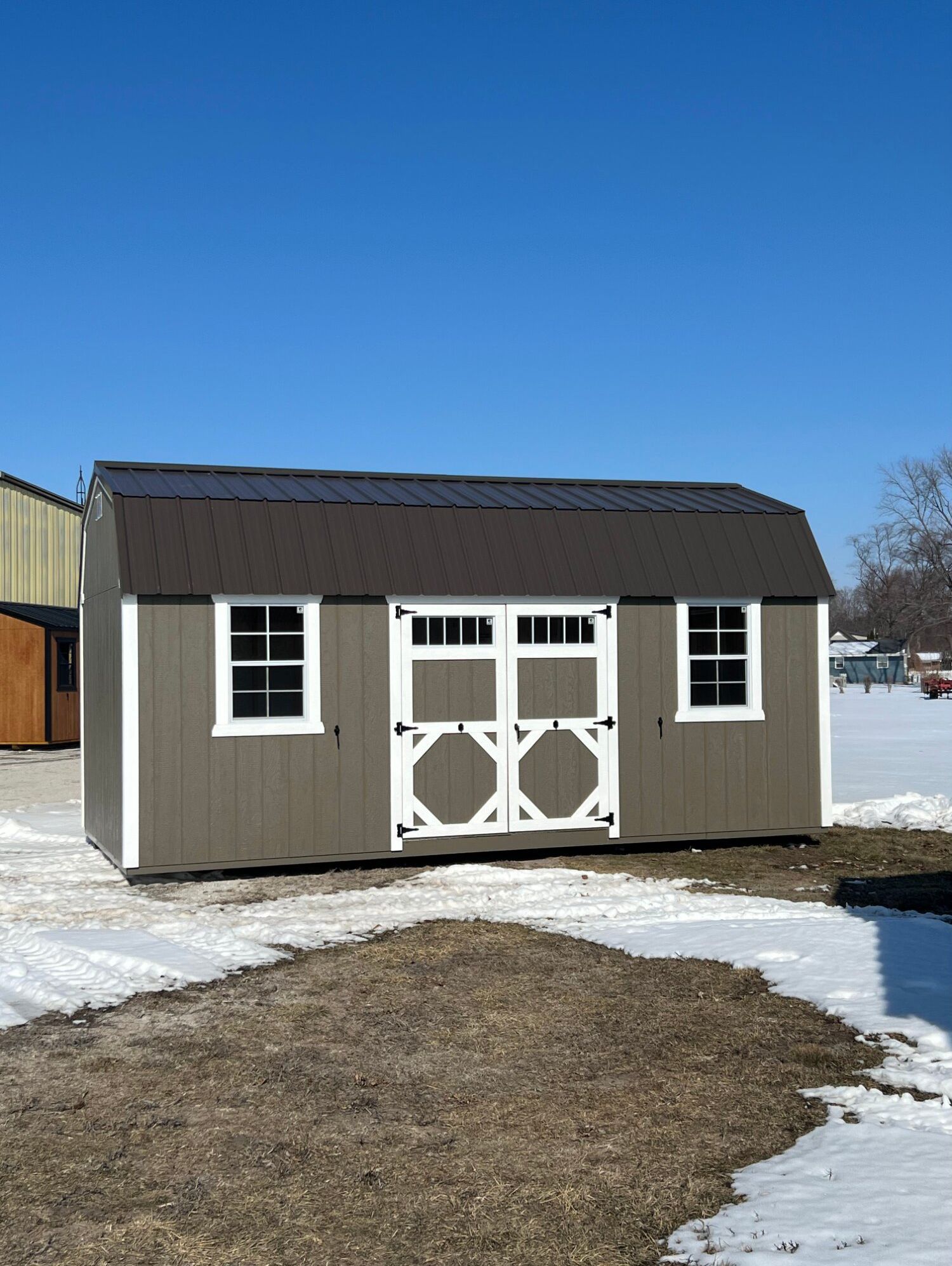 A tan, barn-style shed with white trim, a dark brown metal roof, and double doors, situated on a snowy lot.