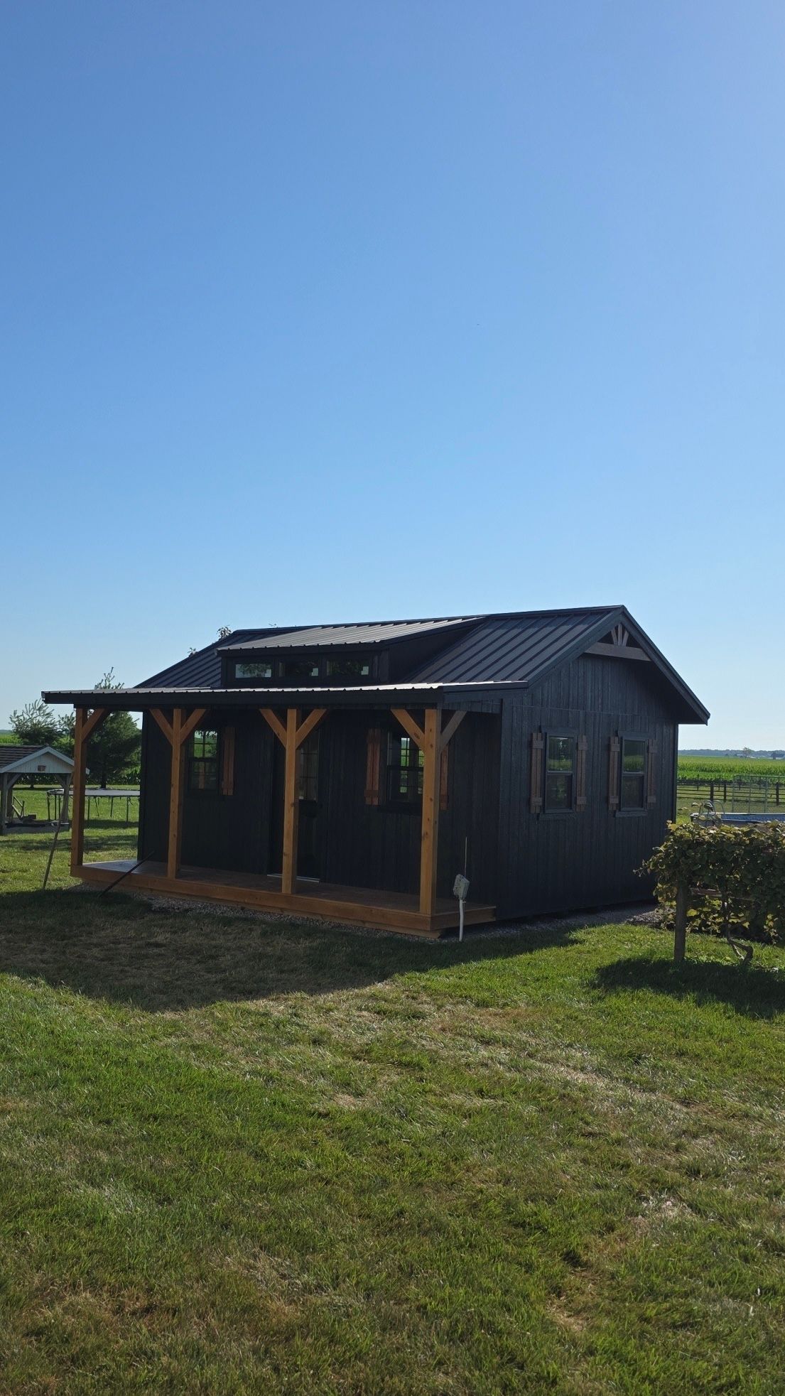 Dark wooden cabin with porch under a blue sky, set in a grassy field.