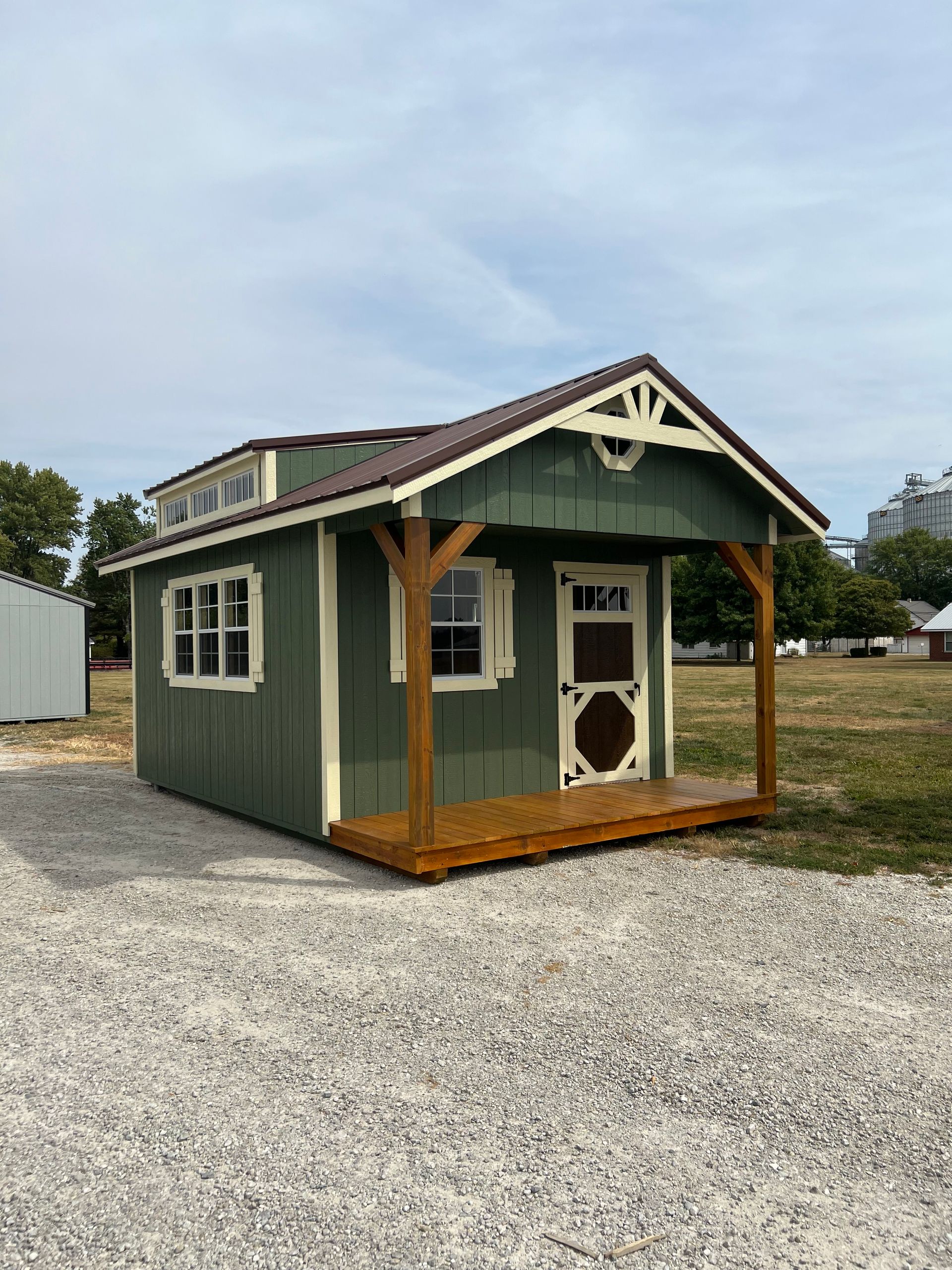 A small green cabin with a front porch, cream trim, and a brown metal roof, sitting on a gravel lot under a cloudy sky.