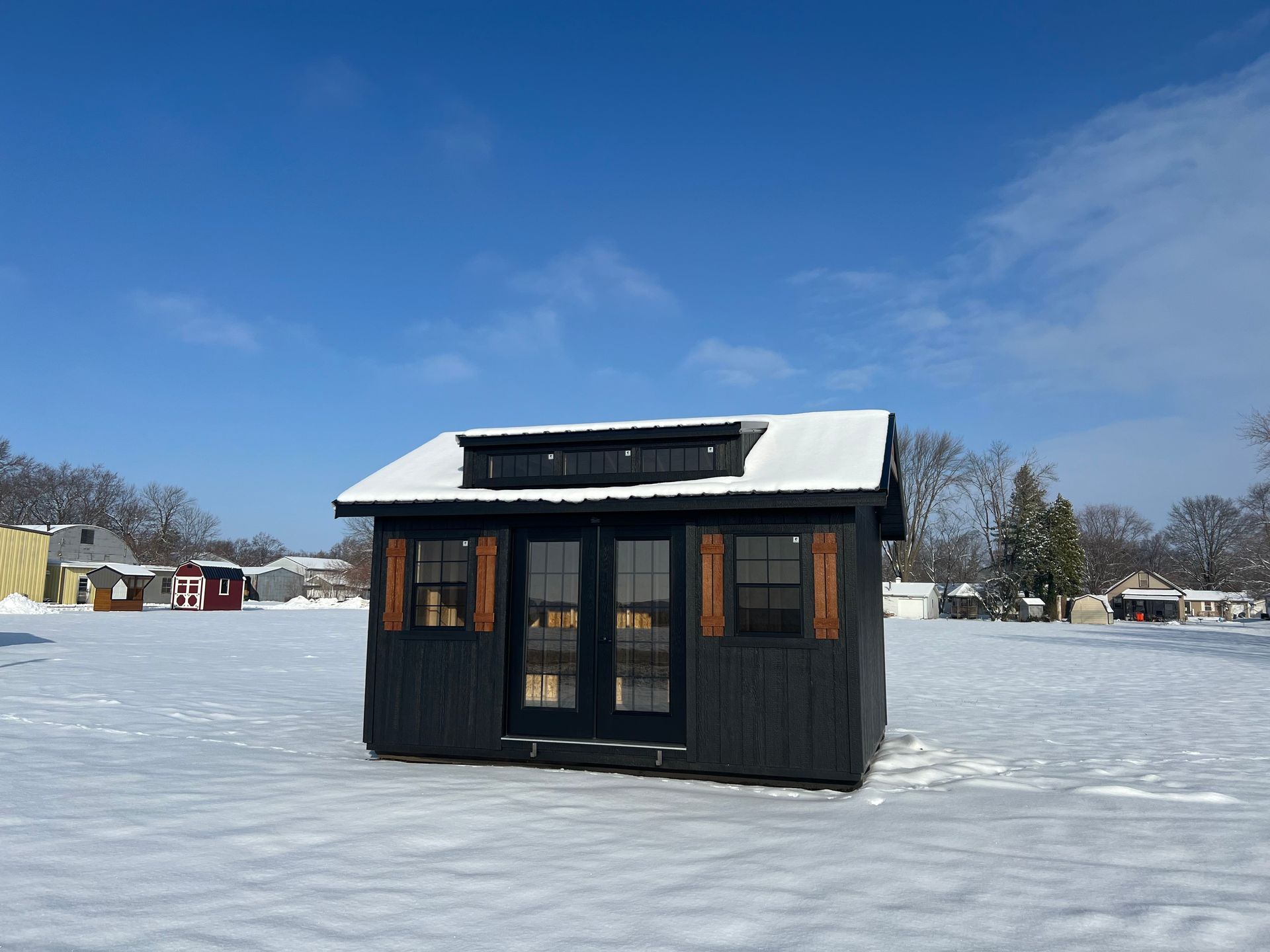 A small, black, modern shed with glass doors and a snowy roof, situated in an open, snow-covered field under a blue sky.