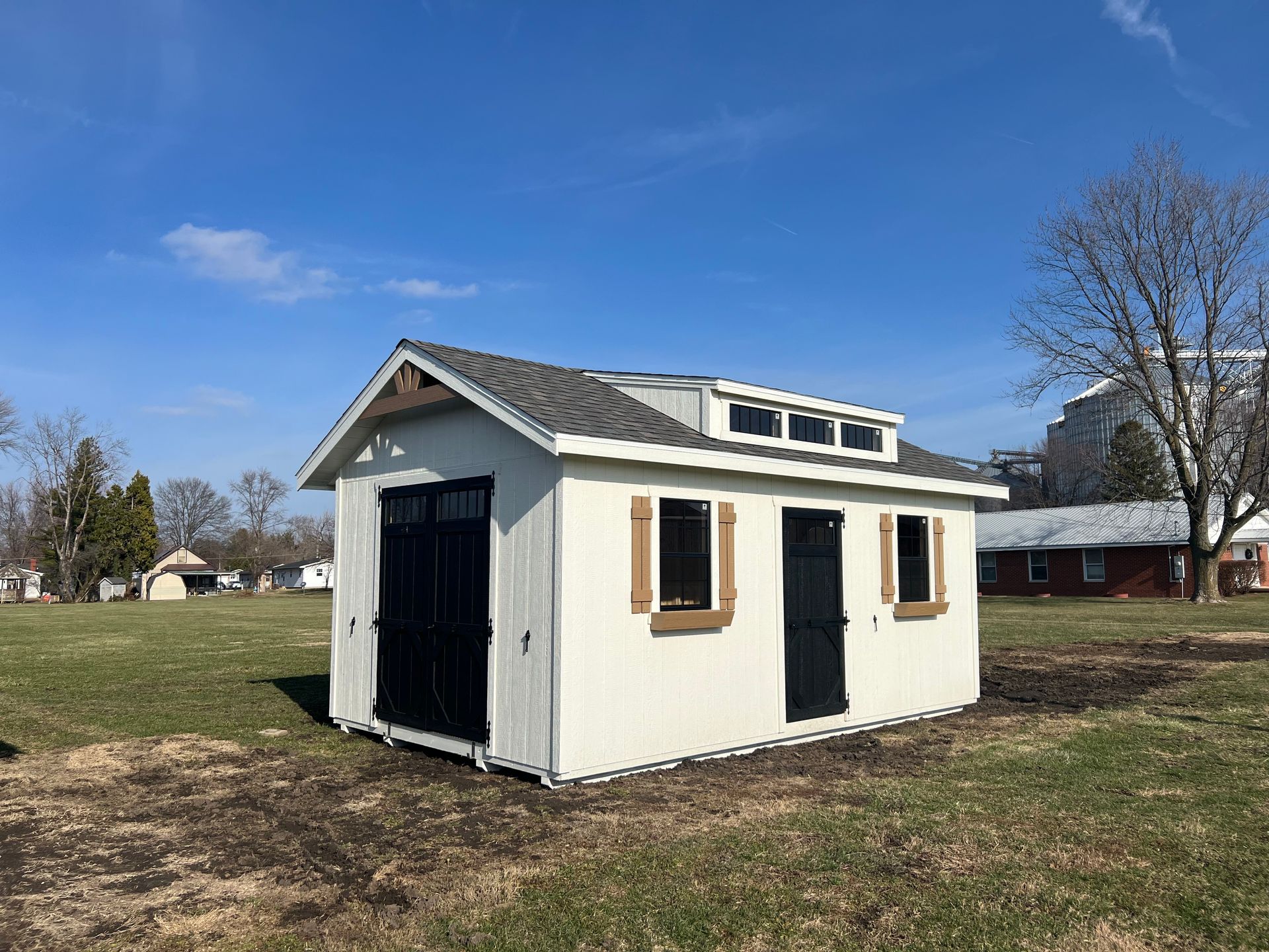 A cream-colored shed with a dark roof and black doors sits in an open, grassy field under a bright blue sky.