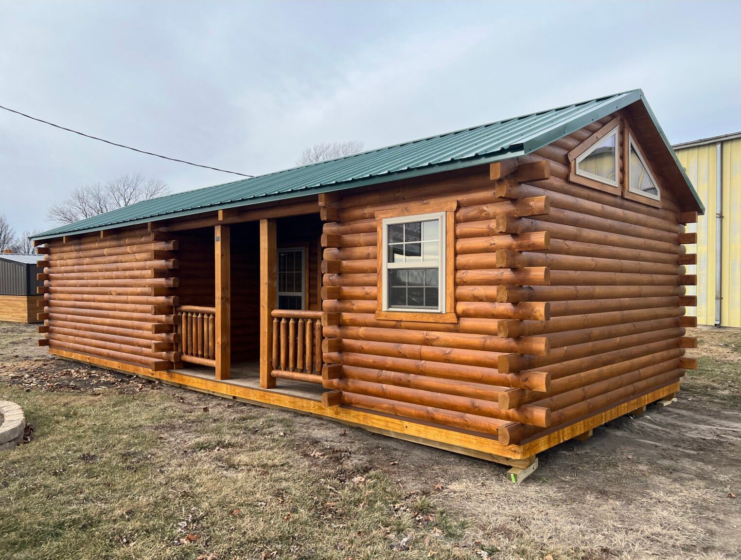 A small log cabin with a green metal roof, a front porch, and wooden walls sits on a dirt lot under a cloudy sky.