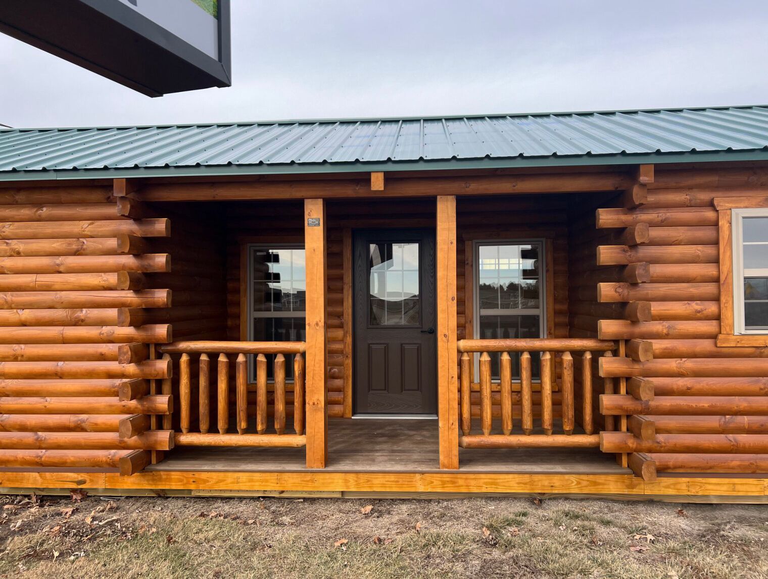 A small, light-brown log cabin with a dark door, windows on each side of the porch, and a green metal roof.