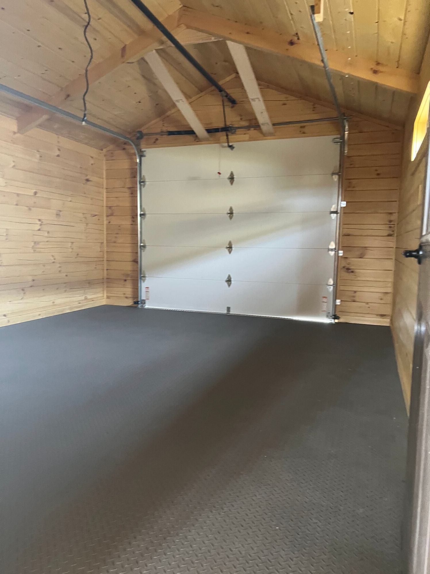 Interior of a wooden garage with light-colored timber walls, a dark grey textured floor, and a closed white garage door.