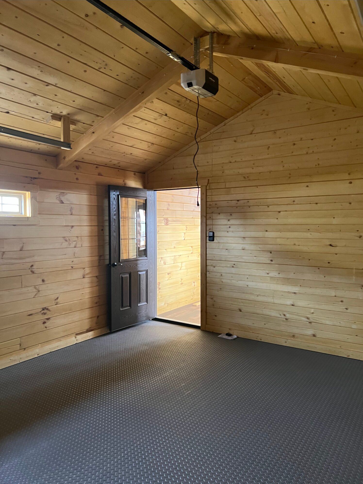 A rustic wood-paneled garage interior with a gray diamond-plate floor, open doorway, and a ceiling-mounted garage opener.