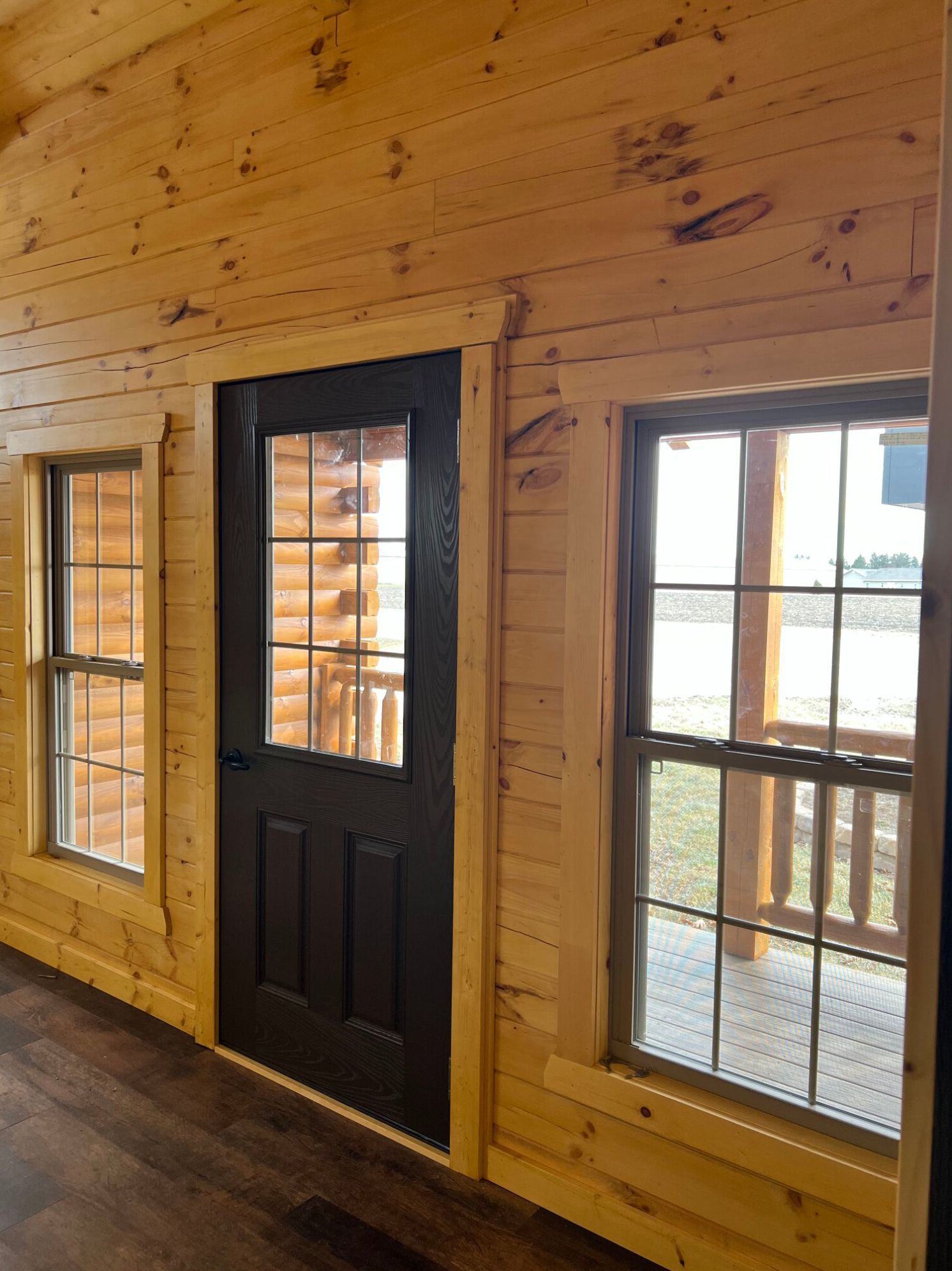 An interior view of a log cabin wall with a dark door flanked by two windows looking out onto a porch.