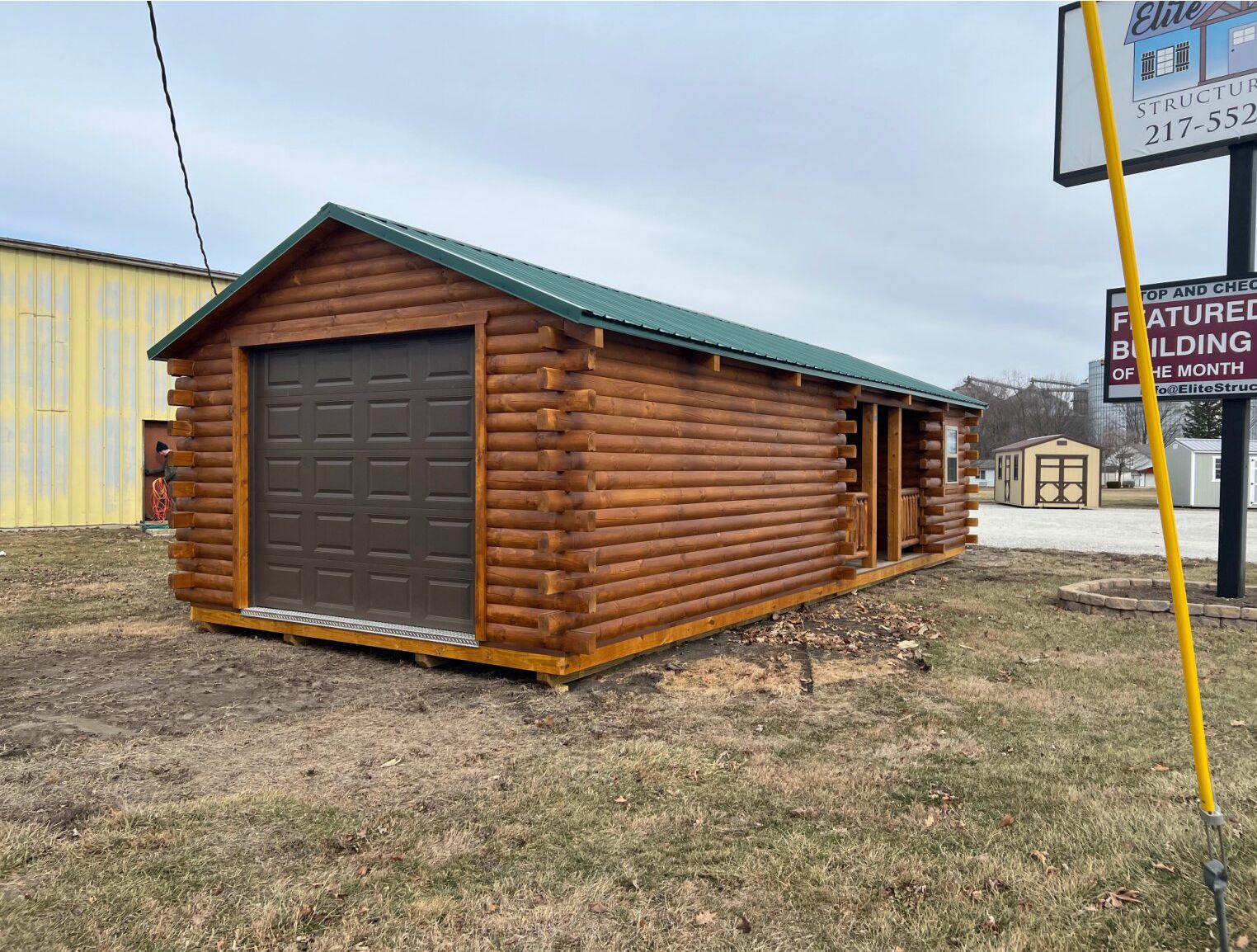 A log-style storage shed with a green metal roof, dark brown garage door, and a side entry on a grassy lot.