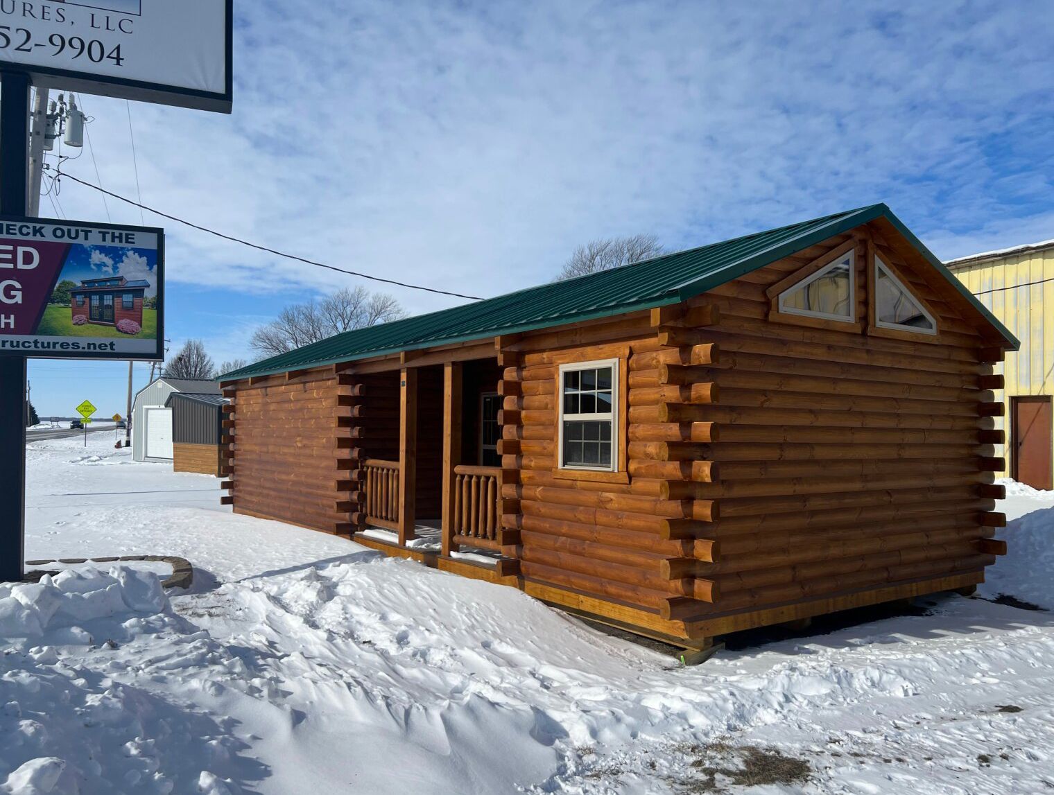 A brown log cabin with a green metal roof and a small front porch sits on a snow-covered lot.